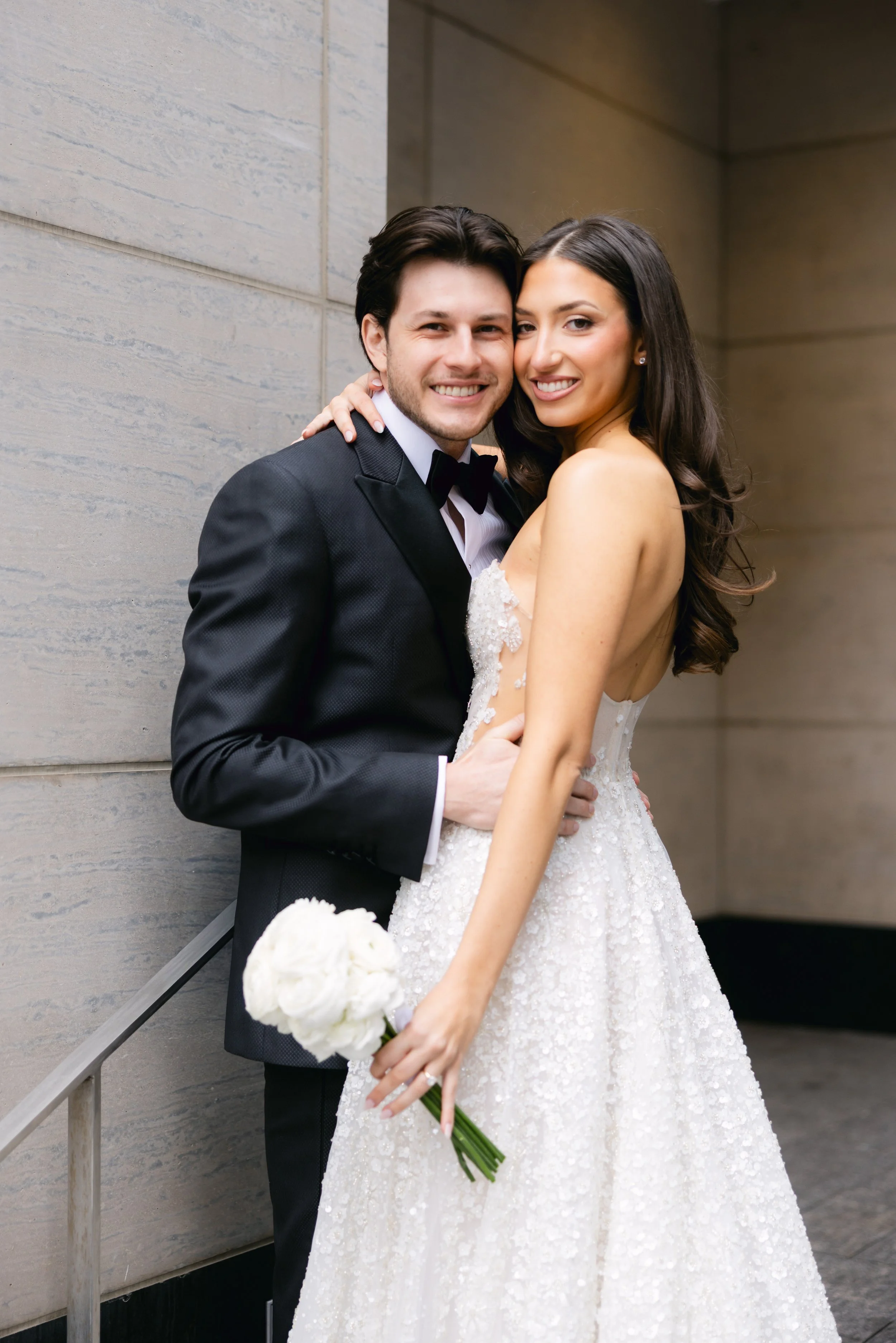 A smiling couple dressed in wedding attire, the man in a black tuxedo and the woman in a white wedding gown holding a bouquet of white flowers, standing closely together.