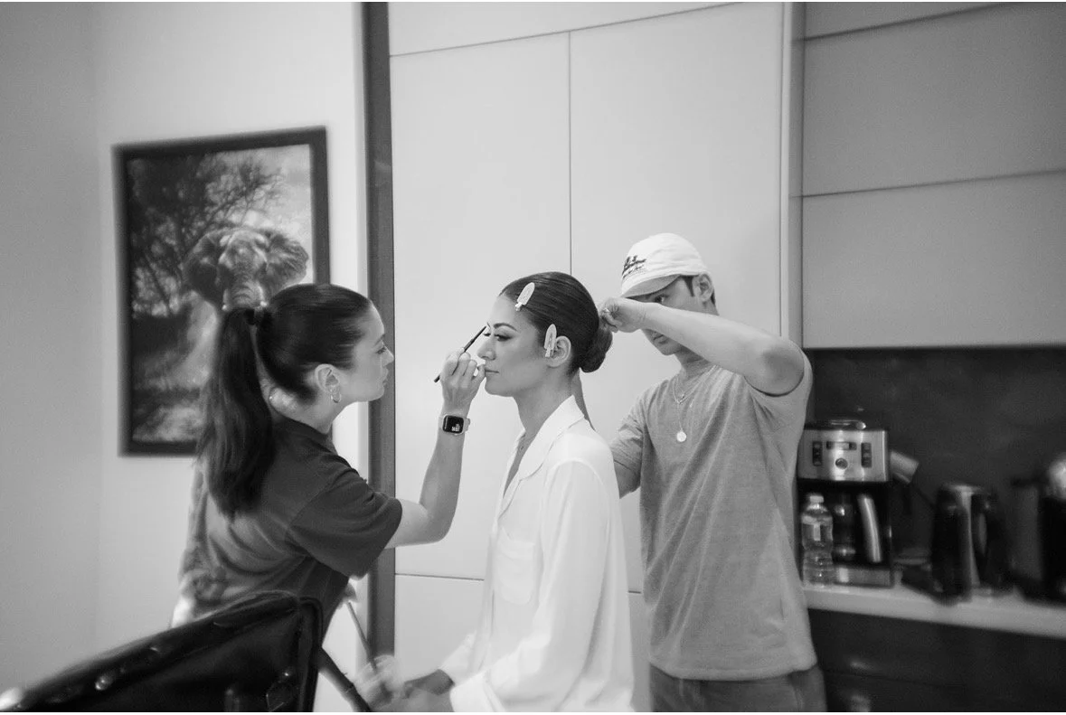 Makeup artist applying makeup to a woman in a white blazer while a man does her hair in a salon or studio setting.