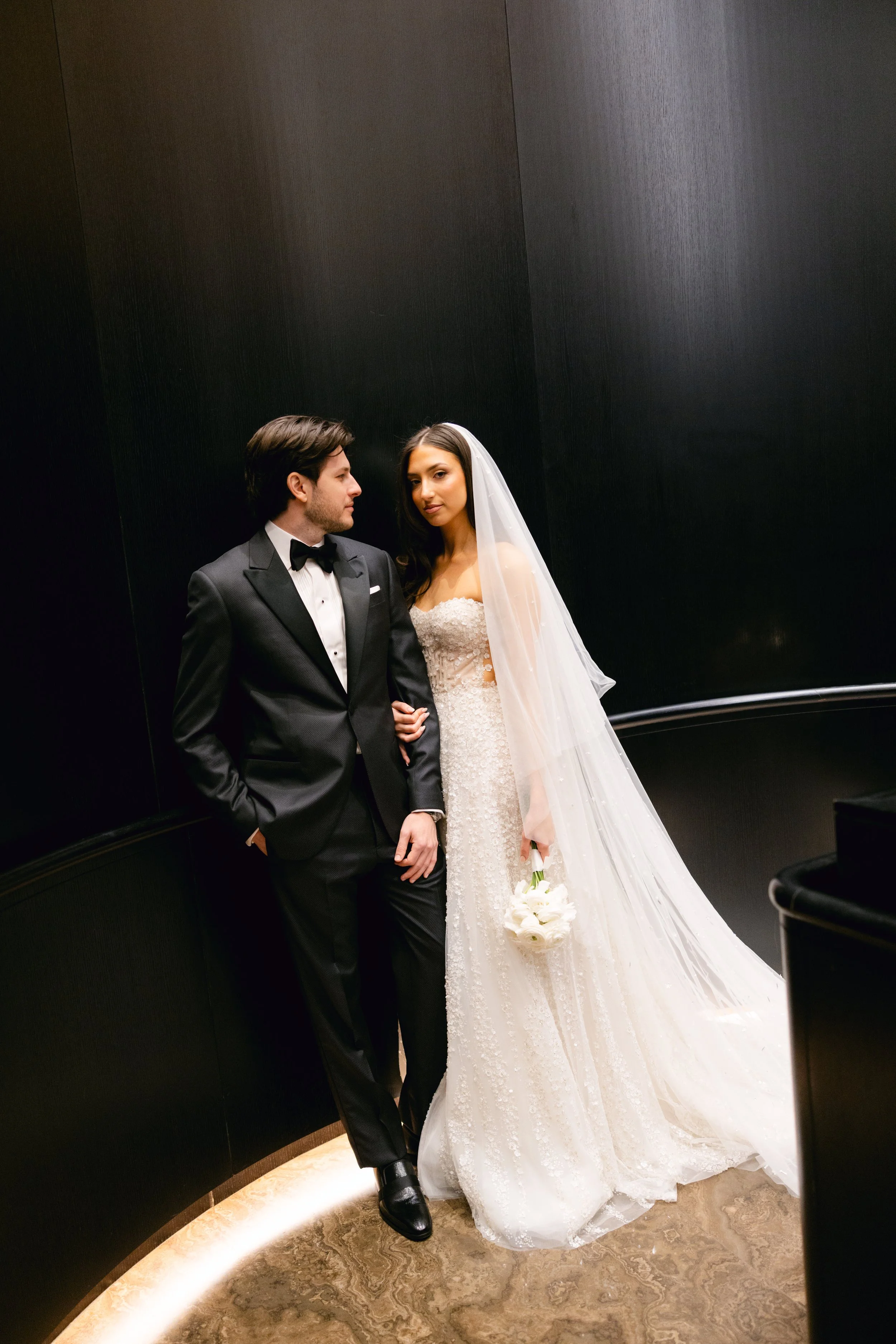 A bride and groom standing together in a dark room, with the groom in a black tuxedo and the bride in a white wedding gown holding a bouquet of white flowers.