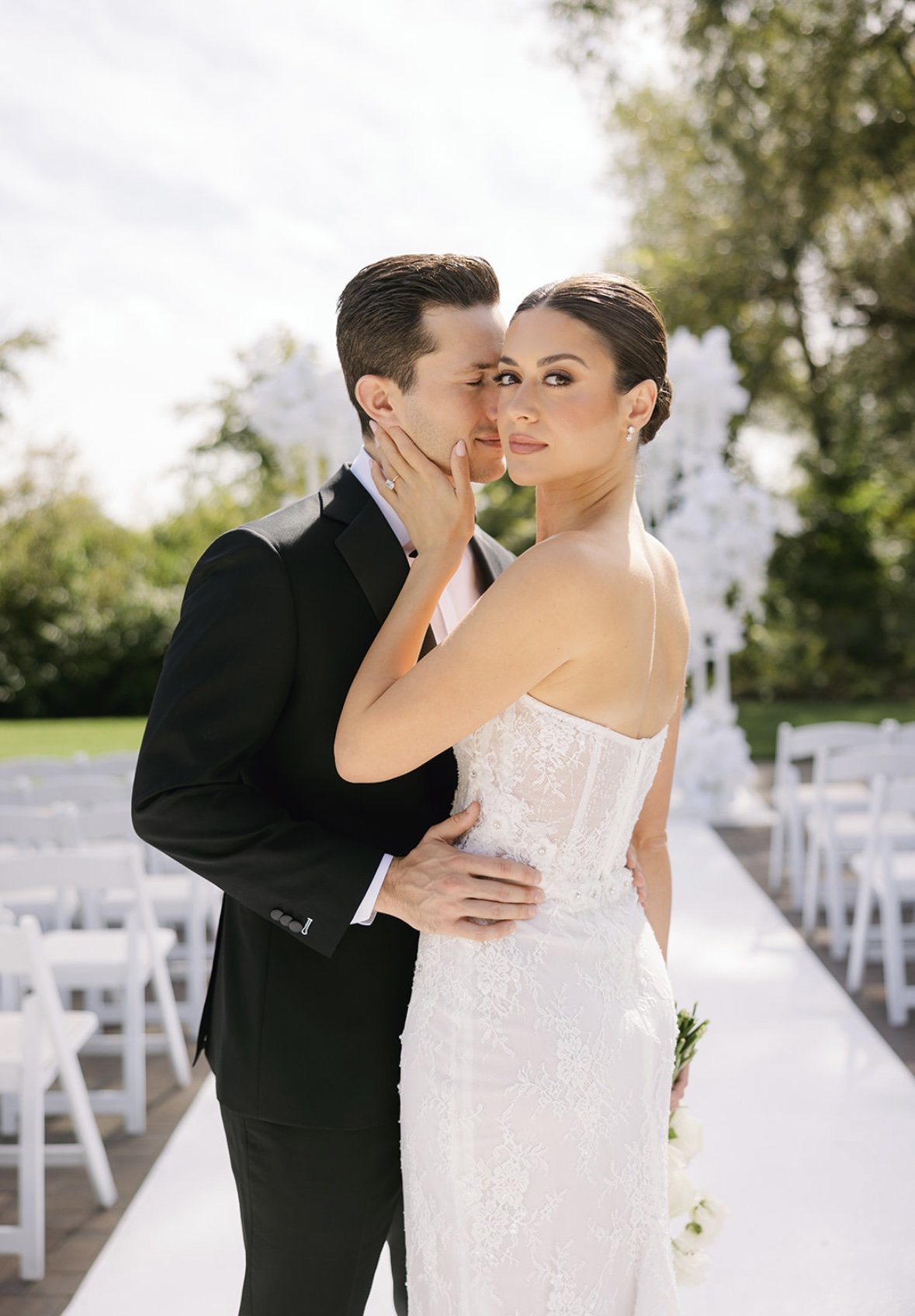A bride and groom in wedding attire sharing an intimate moment outdoors with greenery and white chairs in the background.