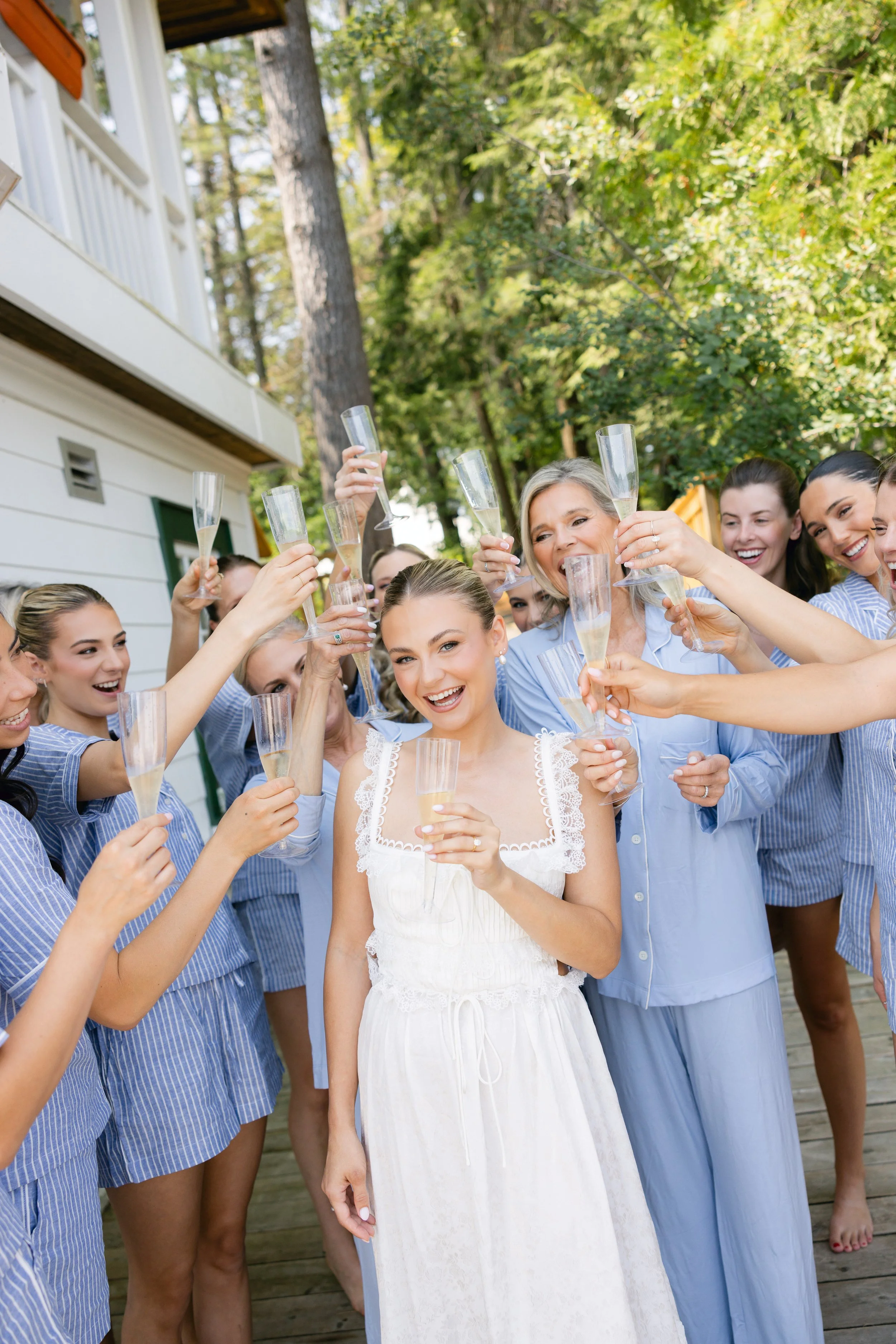 A group of women celebrating outdoors with champagne, some wearing blue pajamas or casual clothing, with green trees in the background.