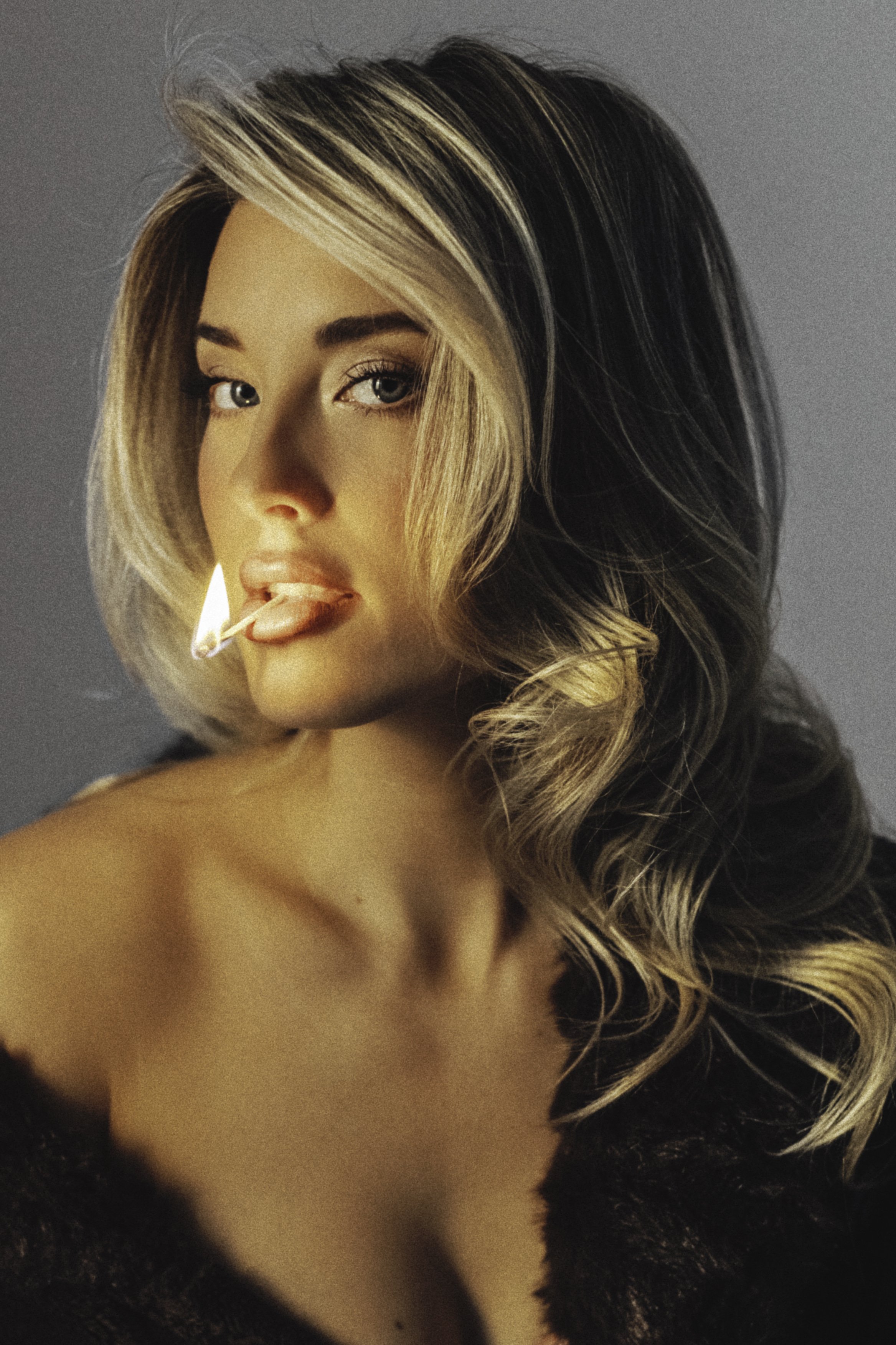 A woman with styled blonde hair, wearing makeup, has a lit matchstick in her mouth, and is dressed in a black furry garment, posing against a neutral background.