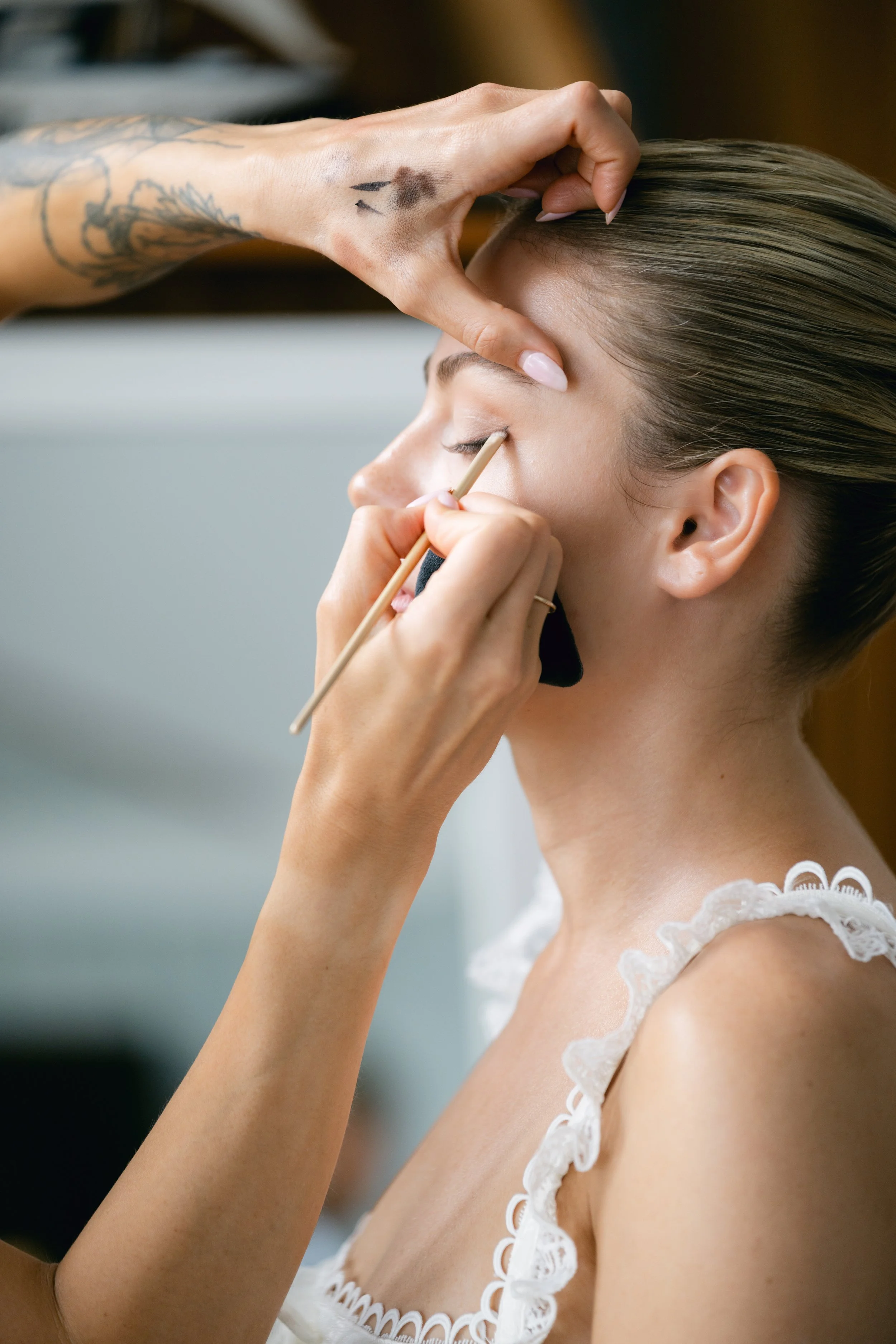 Makeup artist applying eyeshadow to woman with closed eyes, wearing a white lace top, in a beauty salon or studio.