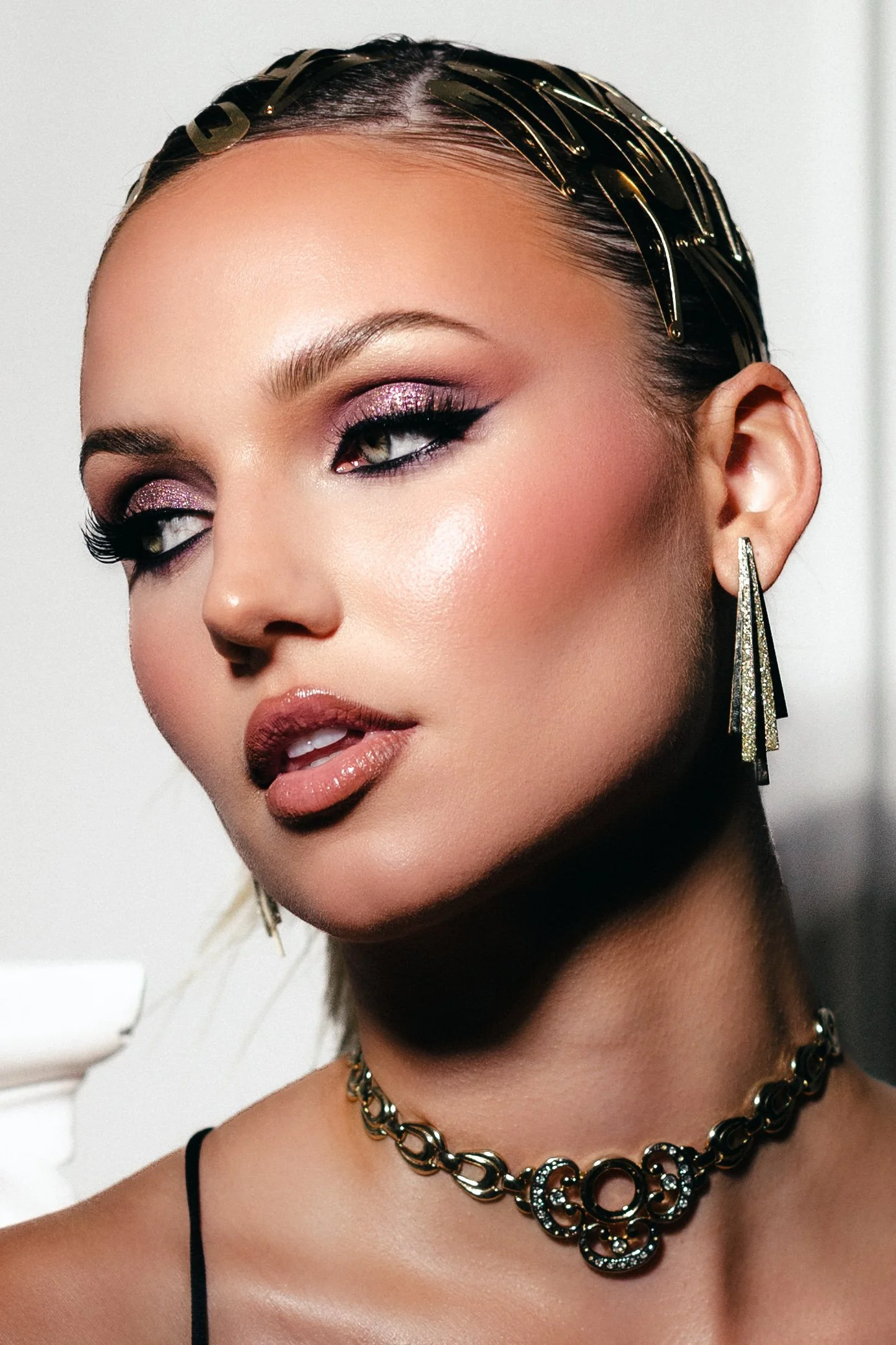 Close-up of a woman with styled hair, bold makeup, glitter eyeshadow, and wearing earrings and a necklace.