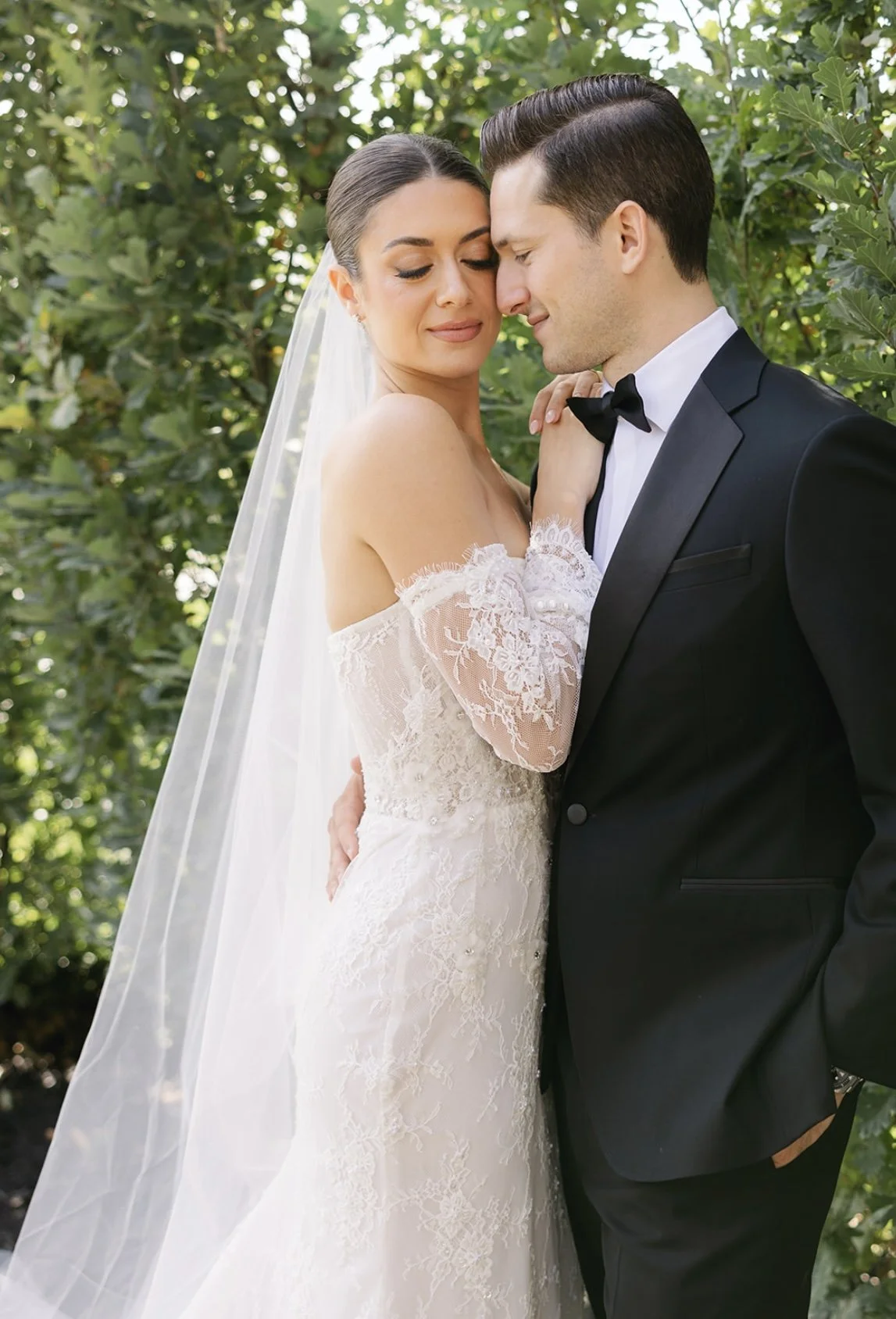 Bride and groom in wedding attire embrace outdoors among greenery, with the bride wearing an off-shoulder lace wedding dress and veil, and the groom in a black tuxedo.