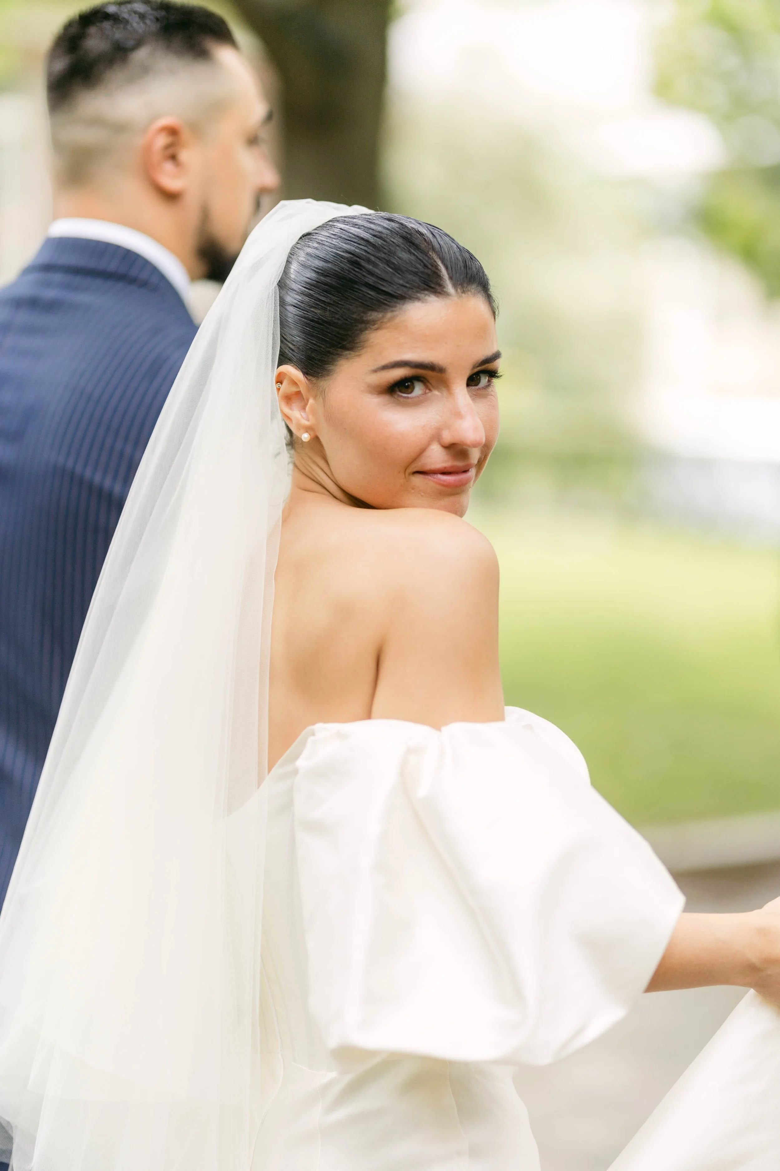 A bride with dark hair and a white off-shoulder wedding dress looking over her shoulder, with a groom in a dark pinstripe suit blurred in the background, outdoors with greenery.