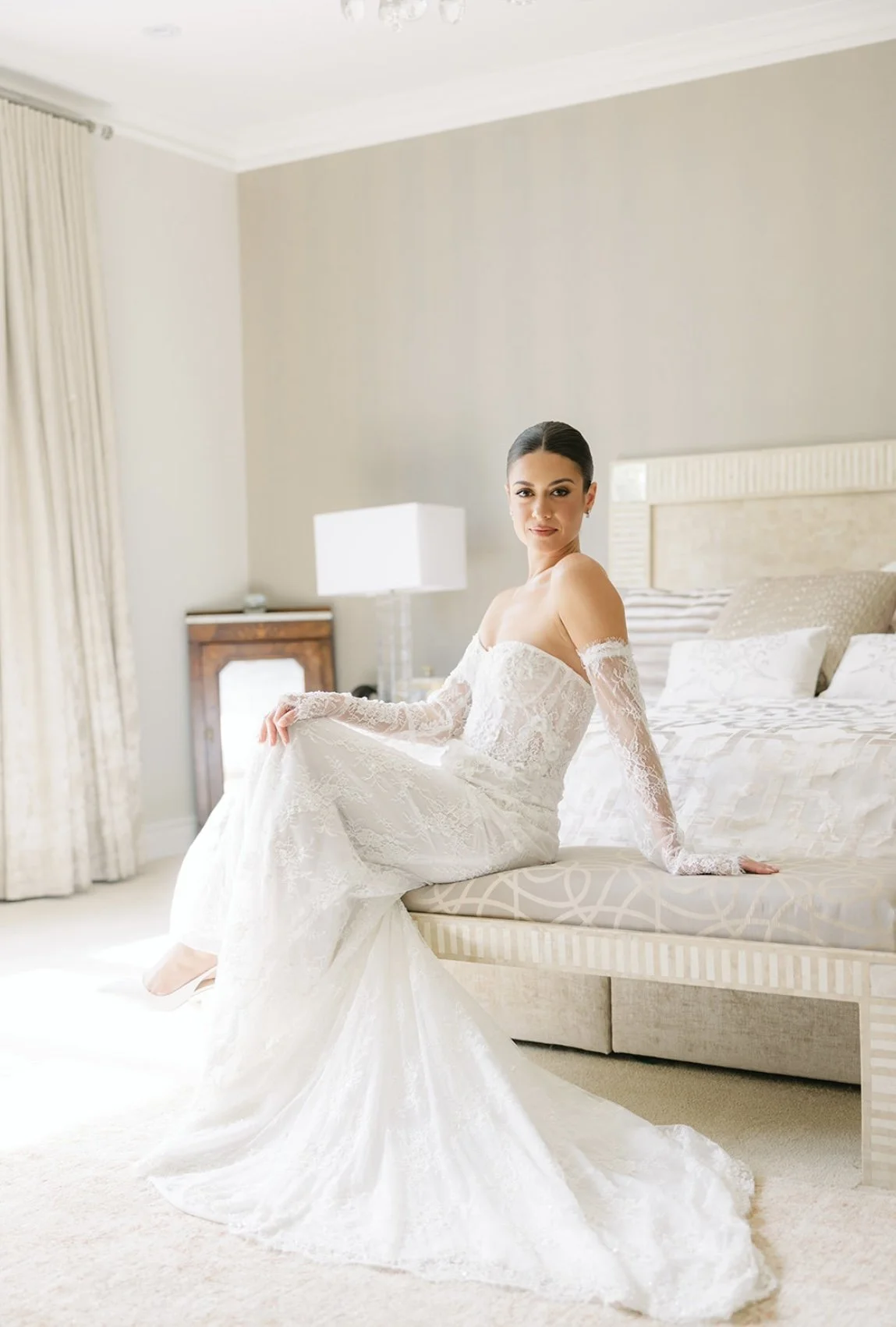 A bride in a white lace wedding dress sitting on the edge of a bed in a well-lit bedroom.
