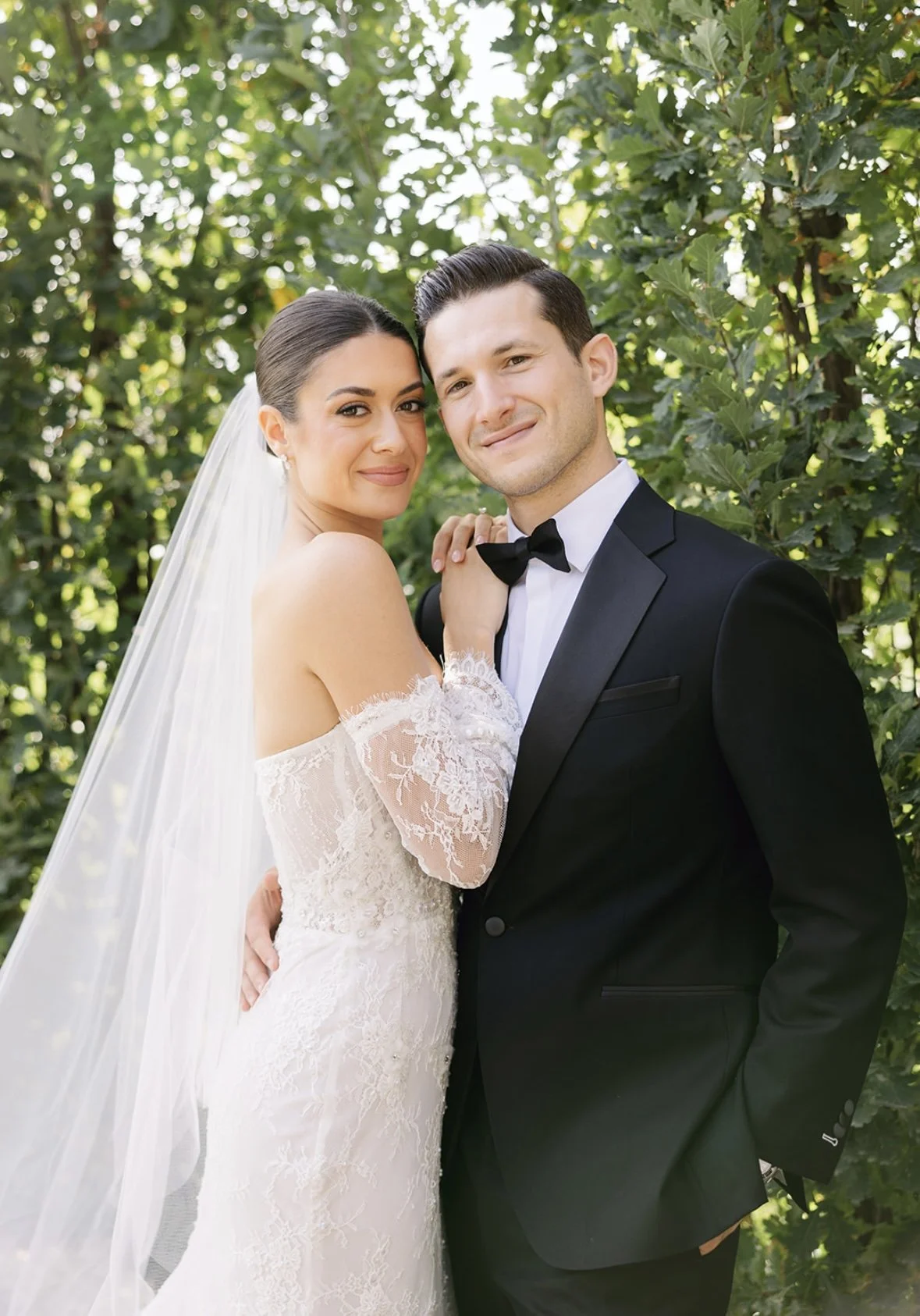 A bride and groom standing close together outdoors with green foliage in the background, dressed in wedding attire, smiling softly at the camera.