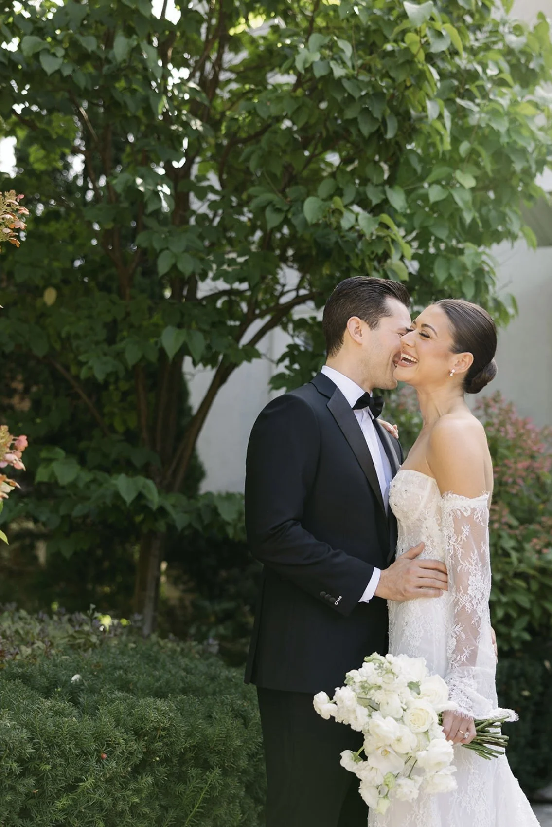 A happy couple dressed in wedding attire sharing a kiss outdoors with greenery in the background. The bride holds a bouquet of white flowers.