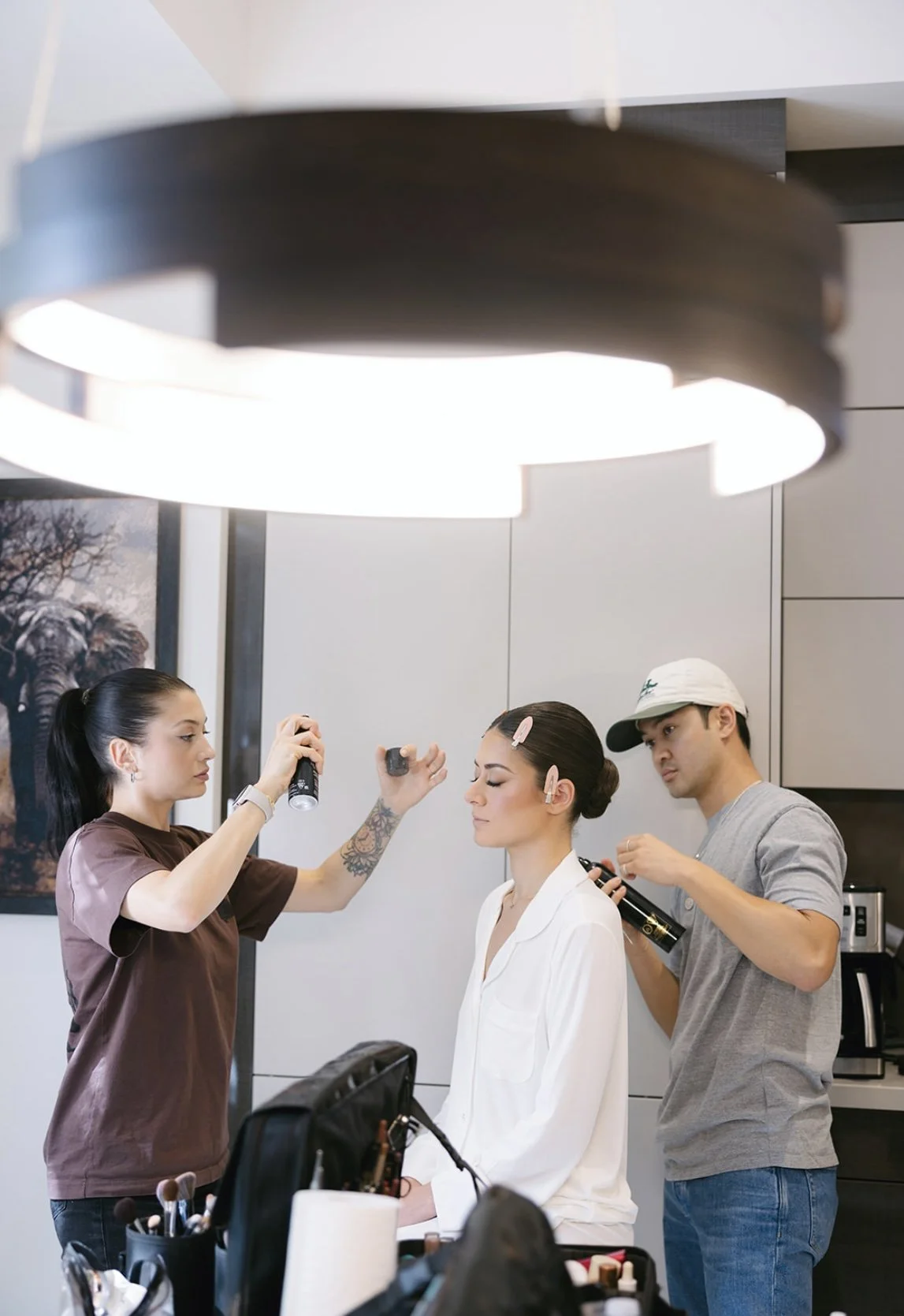 Makeup artists applying makeup to a woman sitting in a white outfit while preparing for a photo or event, with professional makeup tools visible.
