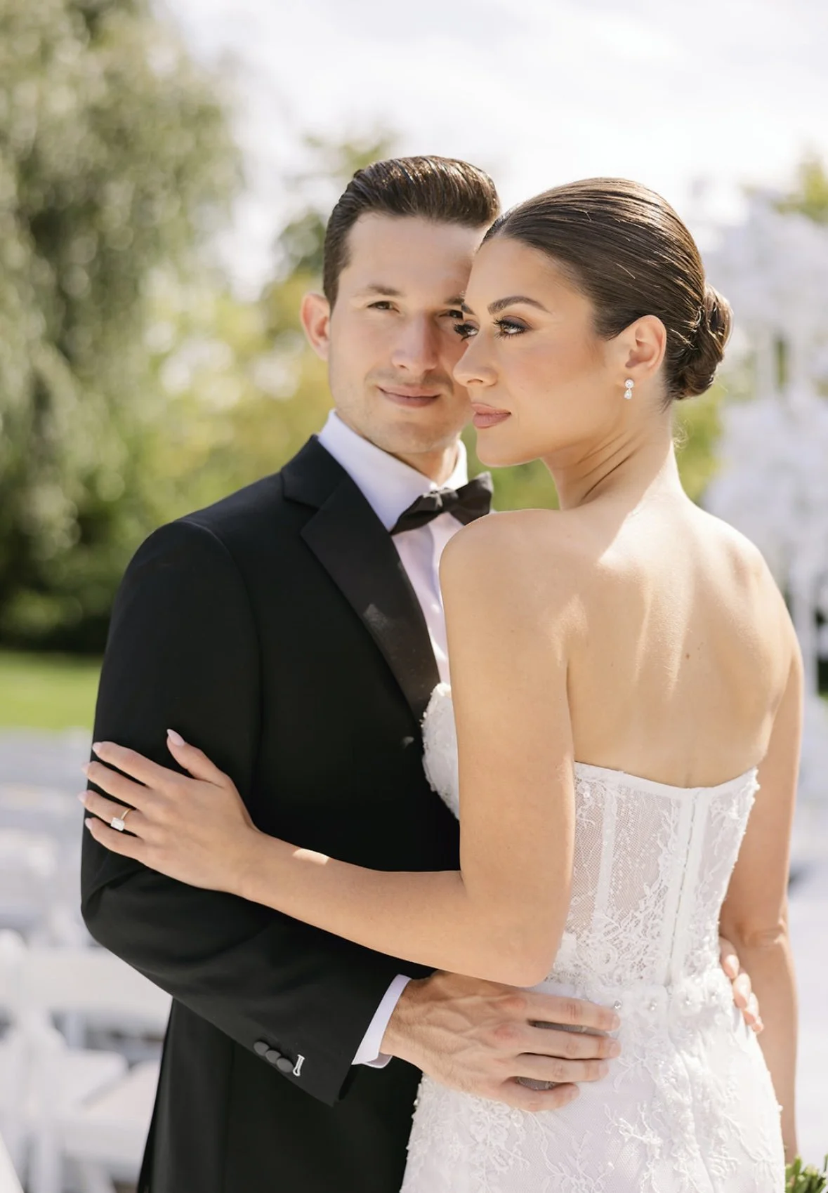 A bride and groom pose outdoors on their wedding day, with the groom wearing a black tuxedo and the bride wearing a strapless white bridal gown. They embrace each other, with the bride's arm around the groom's shoulder and the groom's hand around the
