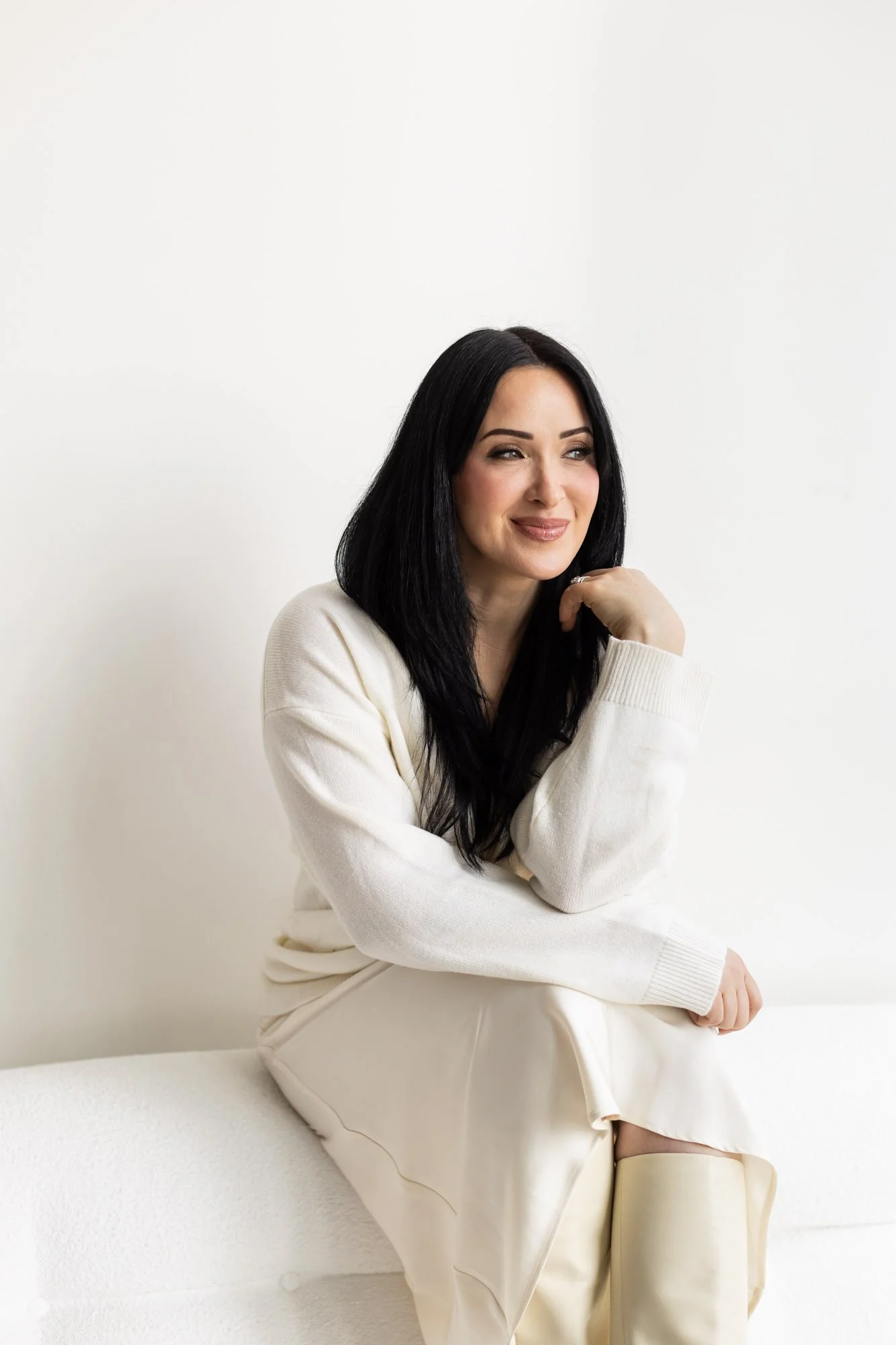 A woman with long black hair and fair skin sits on a white surface against a plain white background. She is wearing a cream-colored sweater and cream-colored pants, and she has a gentle smile while looking slightly to her left.