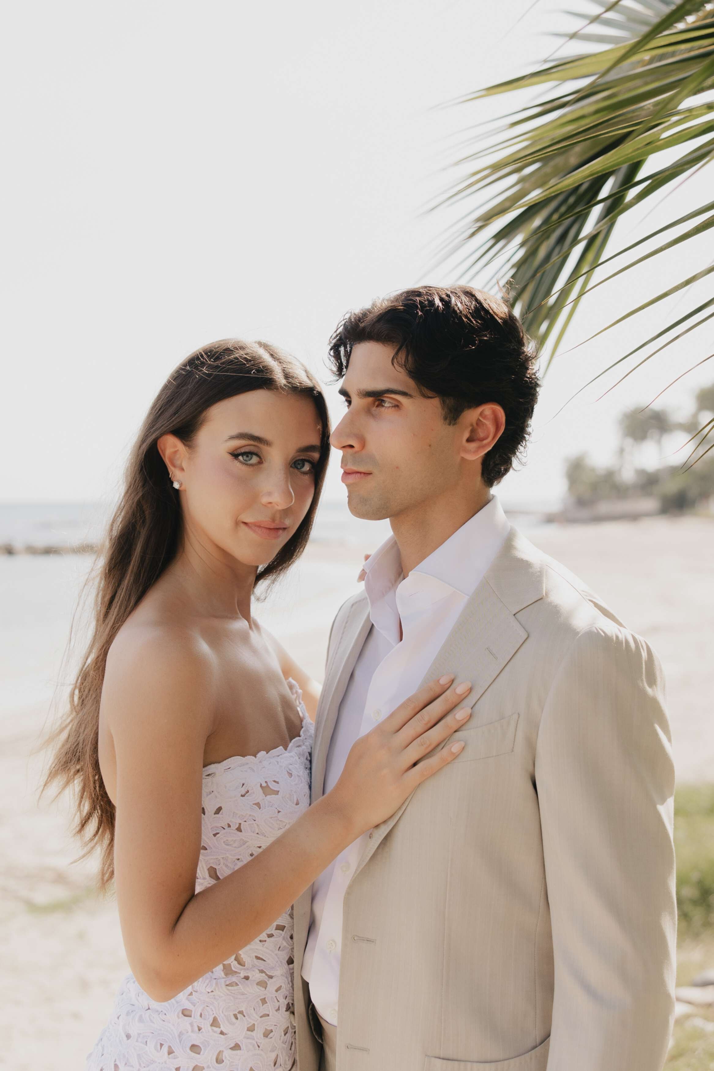 A couple standing closely on a beach, with a palm tree overhead, looking into the camera. The woman has long brown hair and is wearing a white lace dress. The man has dark hair and is dressed in a beige suit with a white shirt.