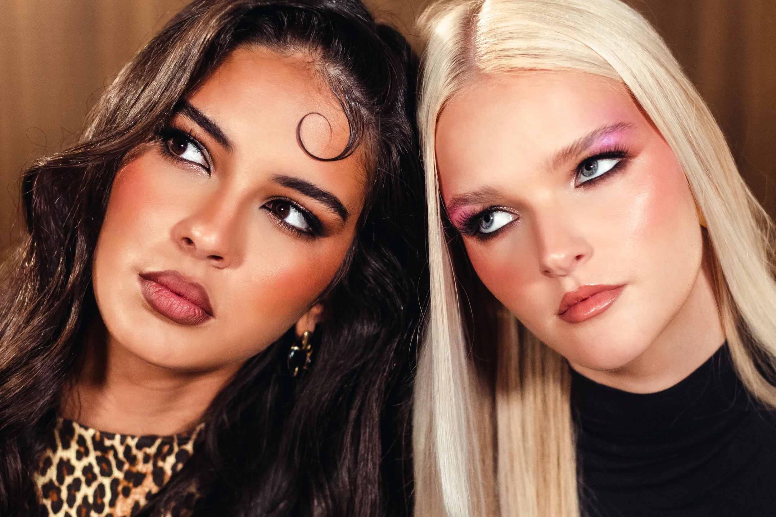 Two women with contrasting hair colors and styles looking to the side, wearing makeup and fashionable clothing.
