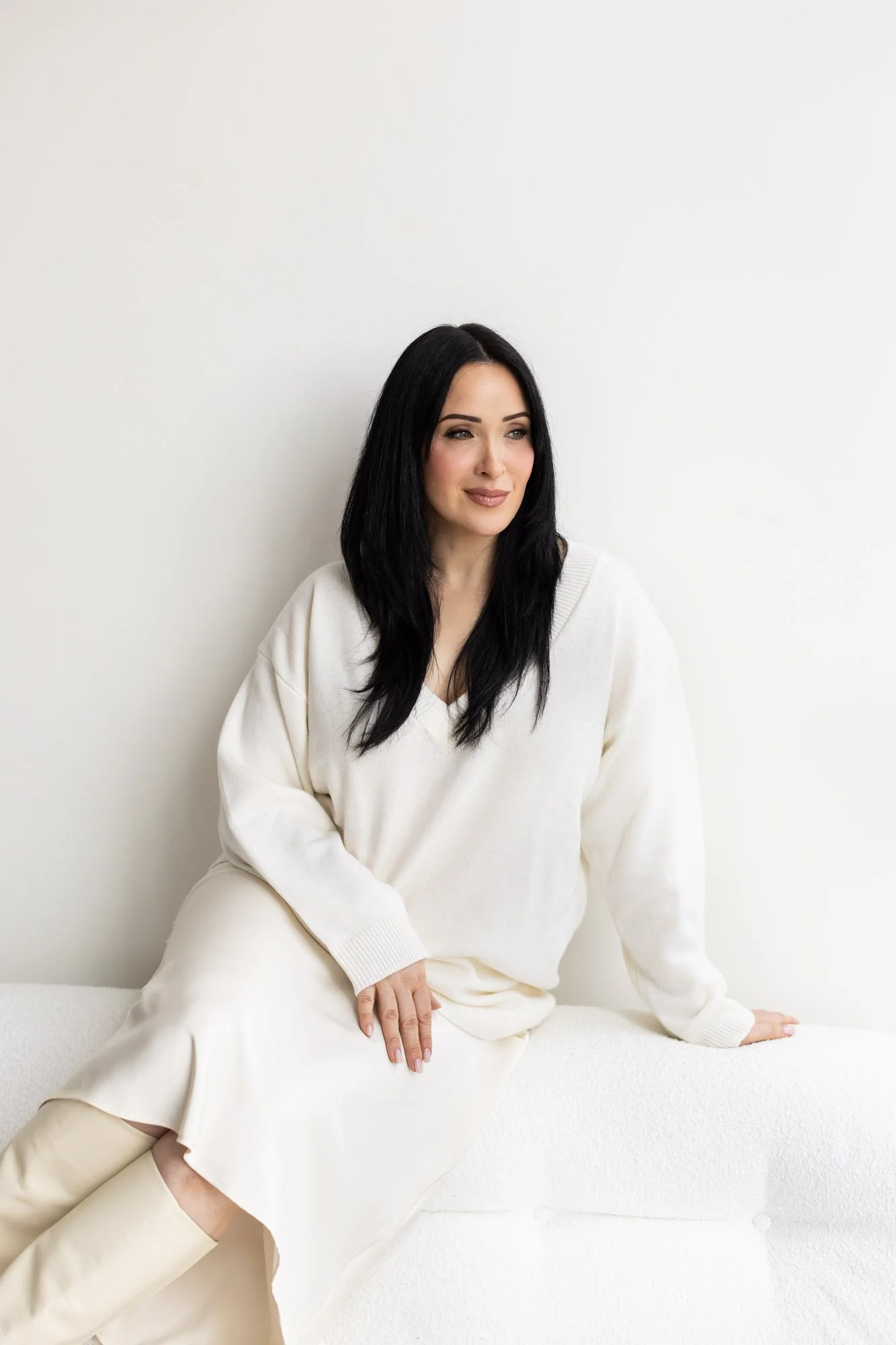 A woman with long black hair and light makeup sitting on a white sofa against a white wall, wearing a cream-colored sweater and skirt.