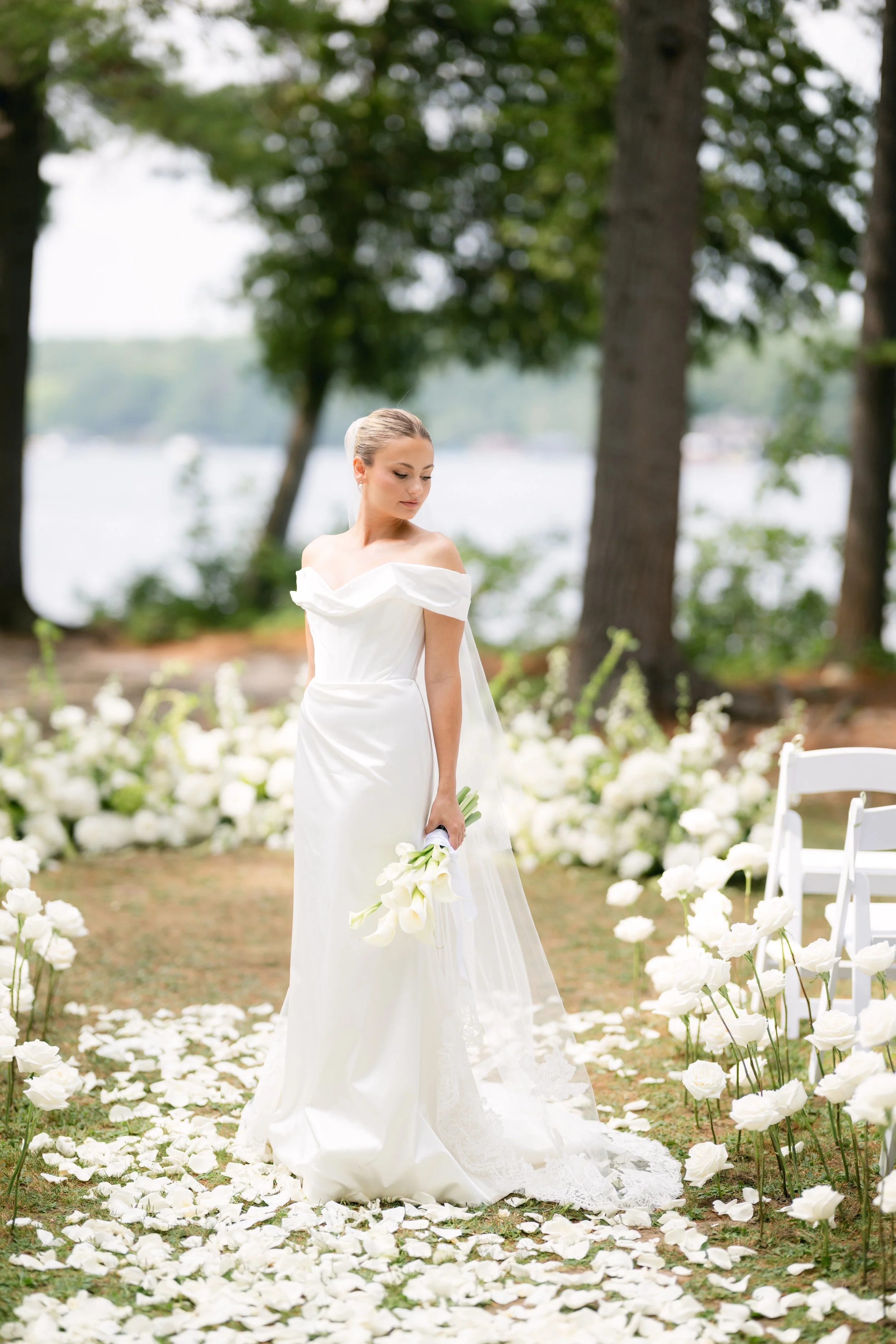 A bride standing outdoors surrounded by white flowers and trees, holding a bouquet of white calla lilies, during a wedding ceremony.