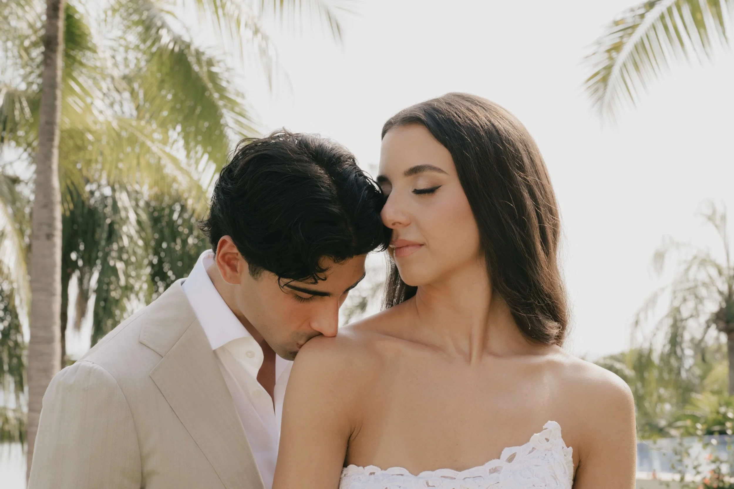 A young man kissing a woman on her shoulder outdoors, with palm trees and bright sky in the background.