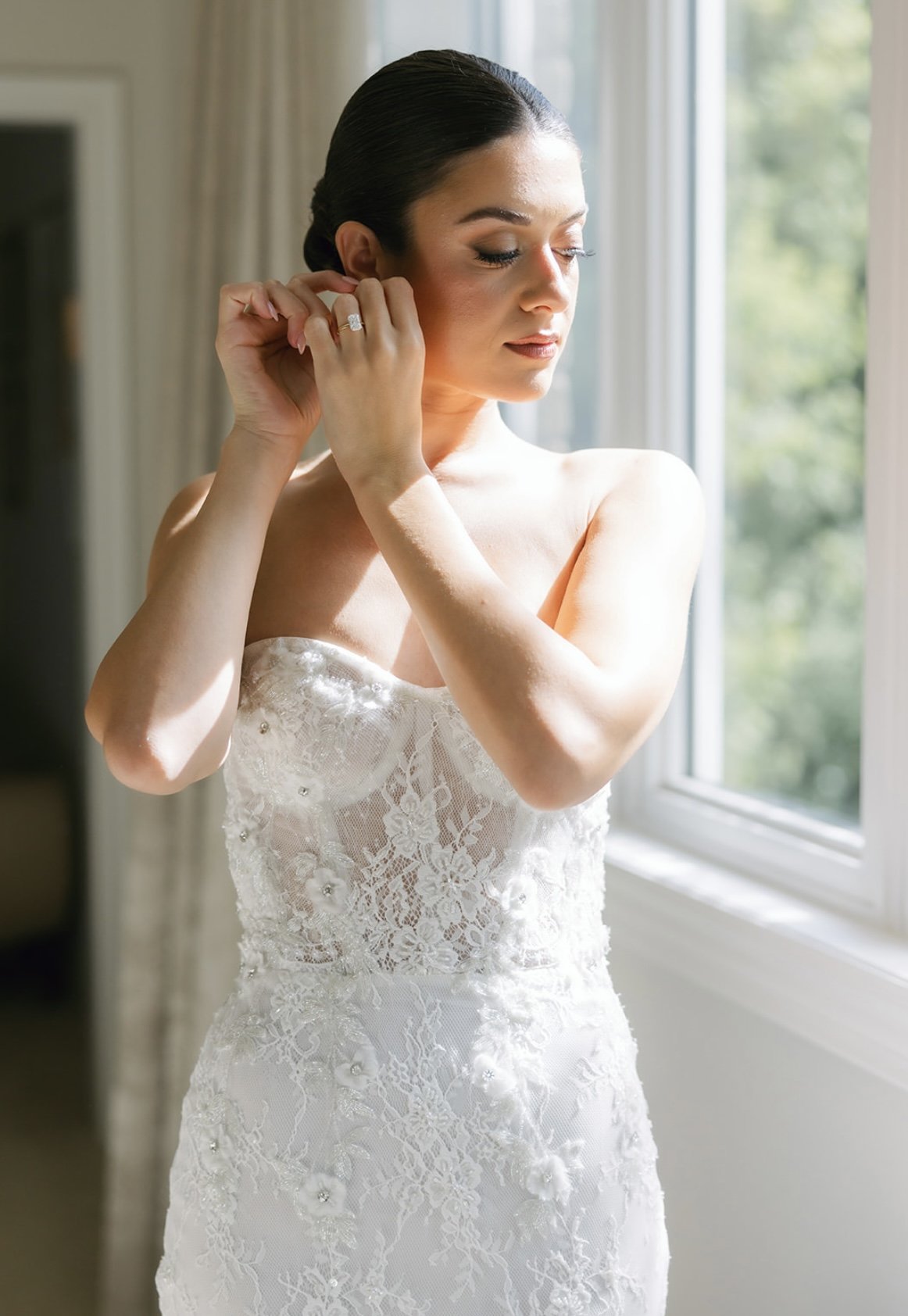 Bride adjusting her earring by a sunlit window, wearing a strapless lace wedding gown with floral embroidery.