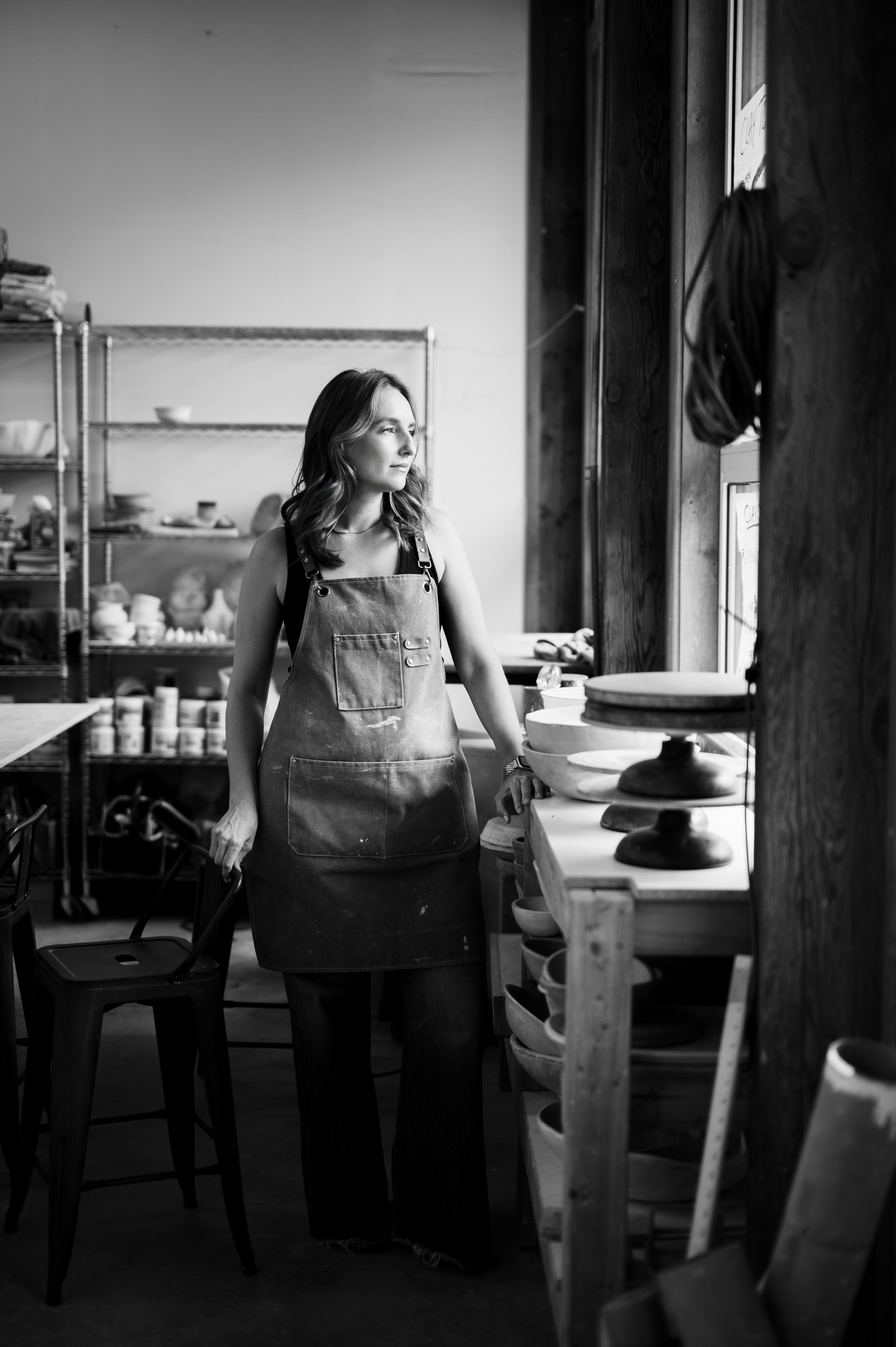 A woman standing in a pottery studio, wearing a apron and looking out the window.