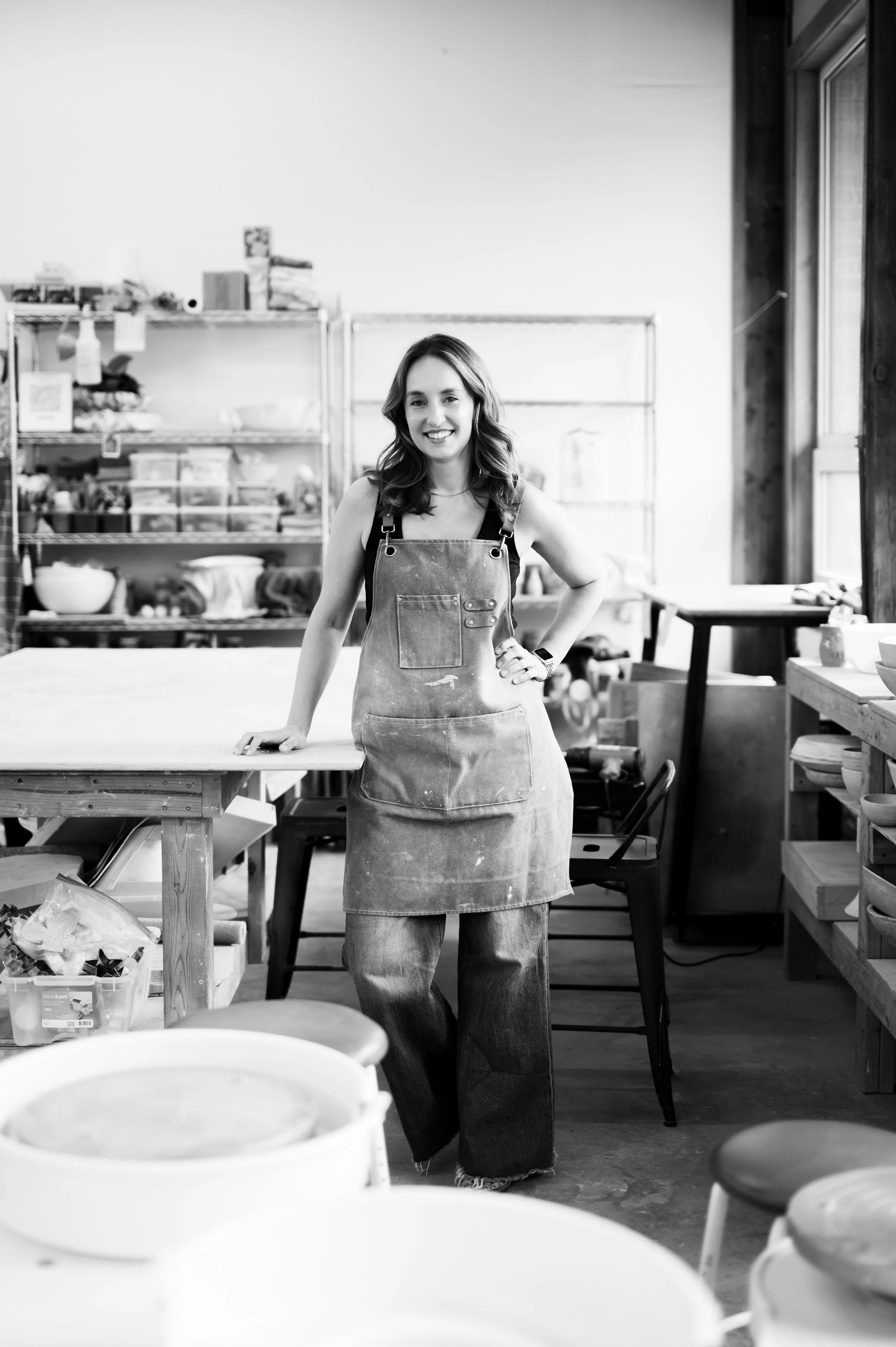 A woman in a workshop or studio, wearing an apron, smiling with her hand on her hip, surrounded by shelves and tables with various supplies and pottery.