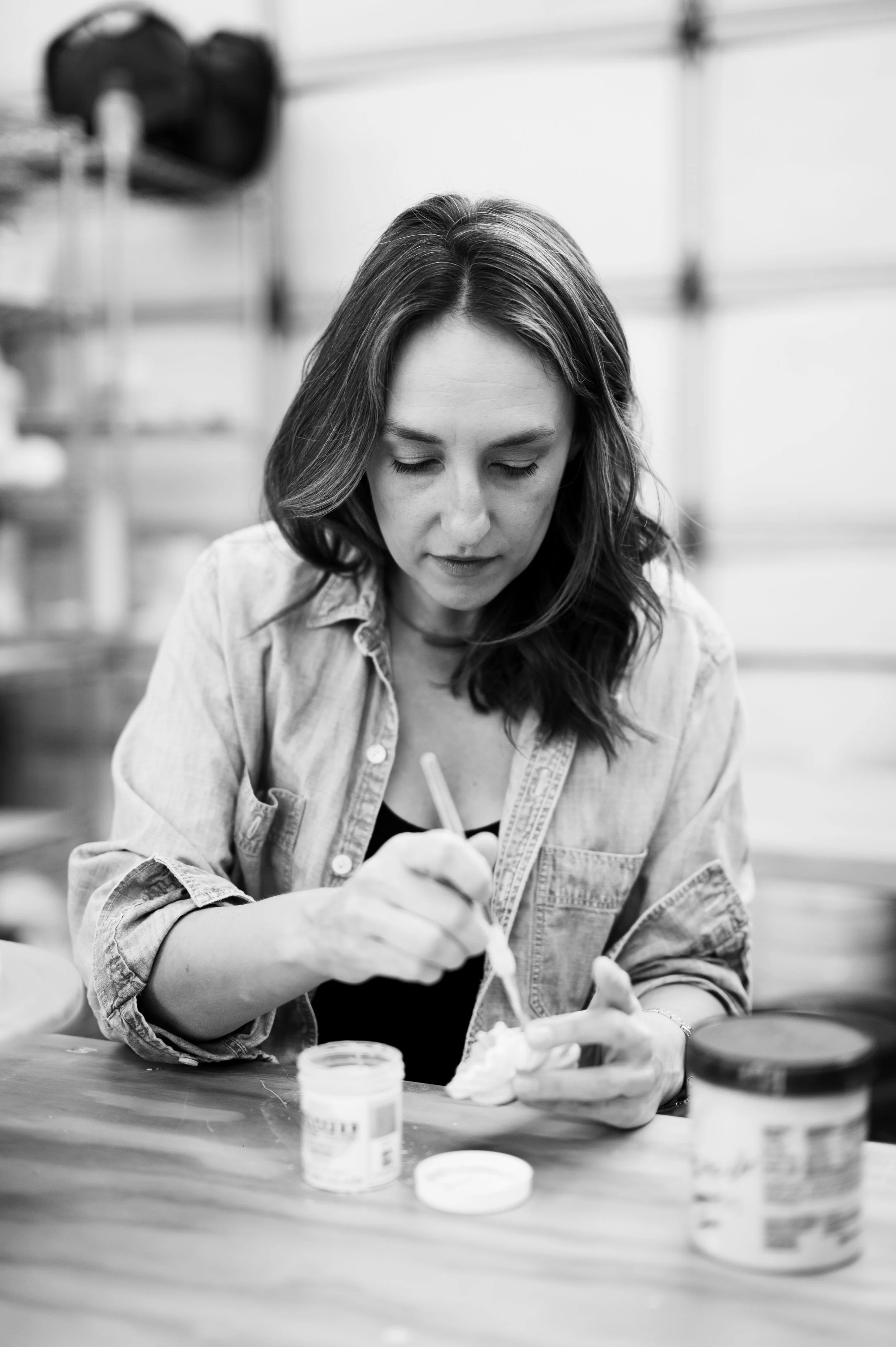 A woman with shoulder-length hair wearing a denim shirt, working with a small container of cream and stirring it with a spatula, in a workshop or laboratory setting.