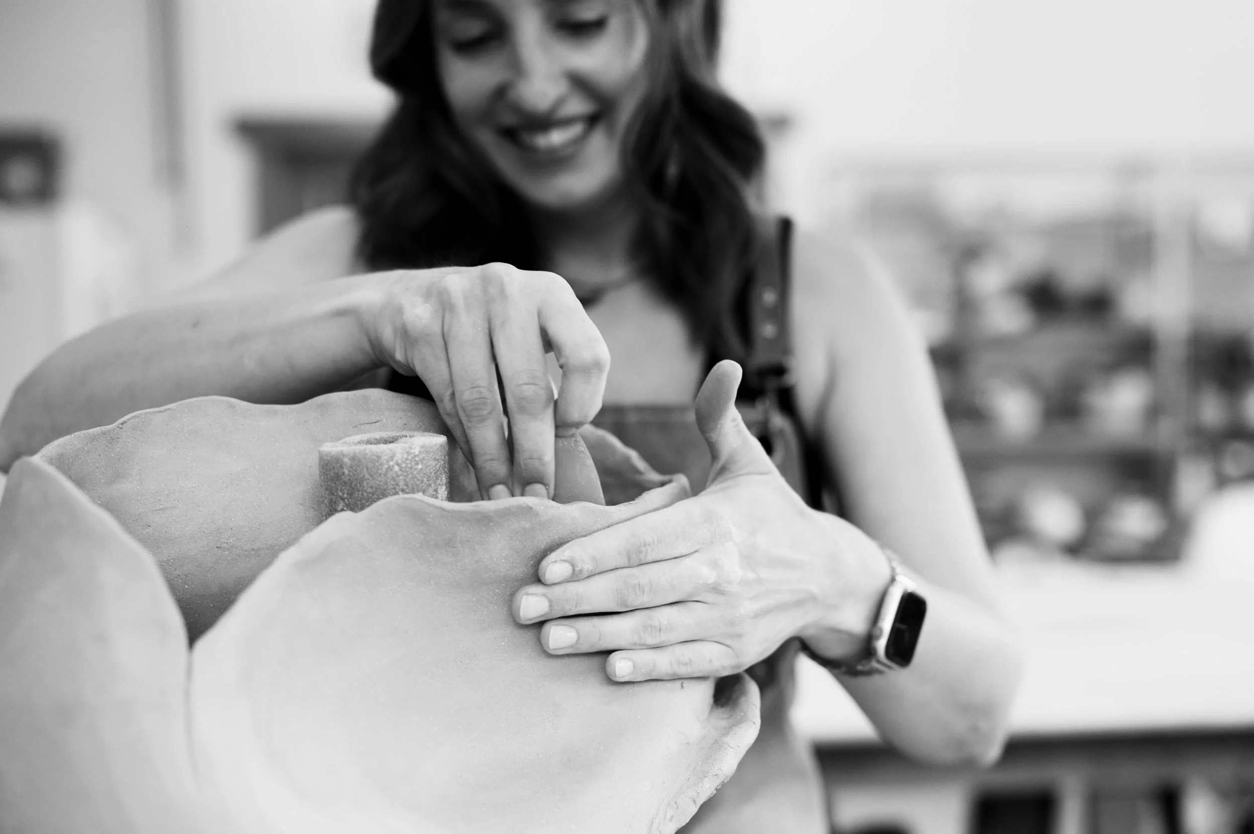 A woman shaping a sculpture in a studio, smiling while working on a clay piece, wearing an apron and a wristwatch.