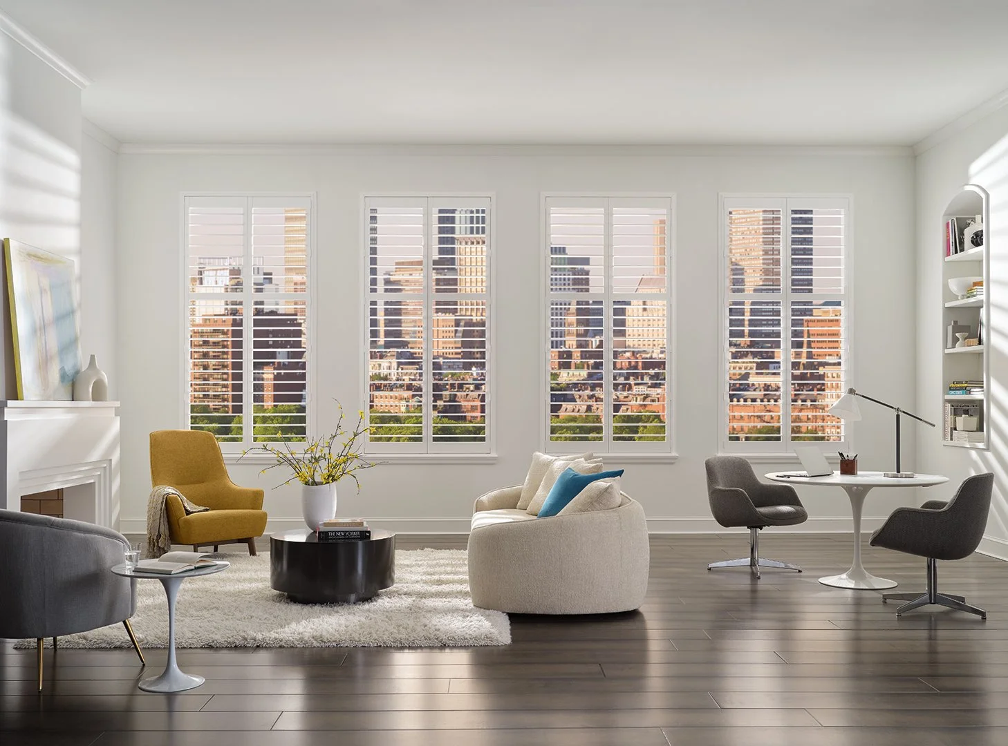 Bright living room with large windows showing city skyline, white walls, dark wood floors, and modern furniture including a yellow armchair, gray chairs, a white sofa with blue cushions, a white round table, a black coffee table with a vase of yellow