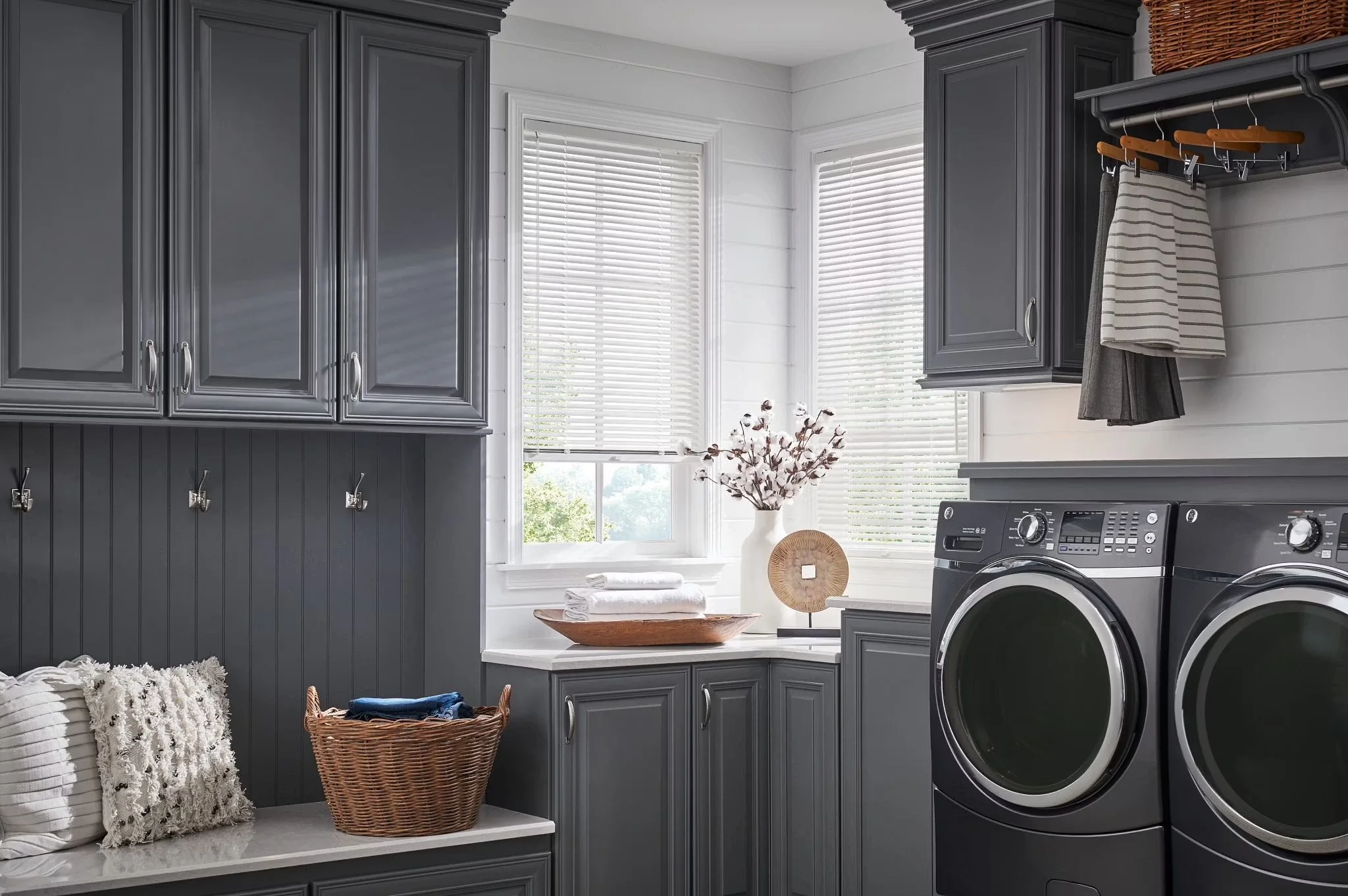 Laundry room with dark gray cabinets, white walls, and a window with blinds. On the counter, there are folded towels, a paper bag with laundry, and a decorative vase with branches. A washing machine is on the right side. There is a basket with laundry and towels on the floor.