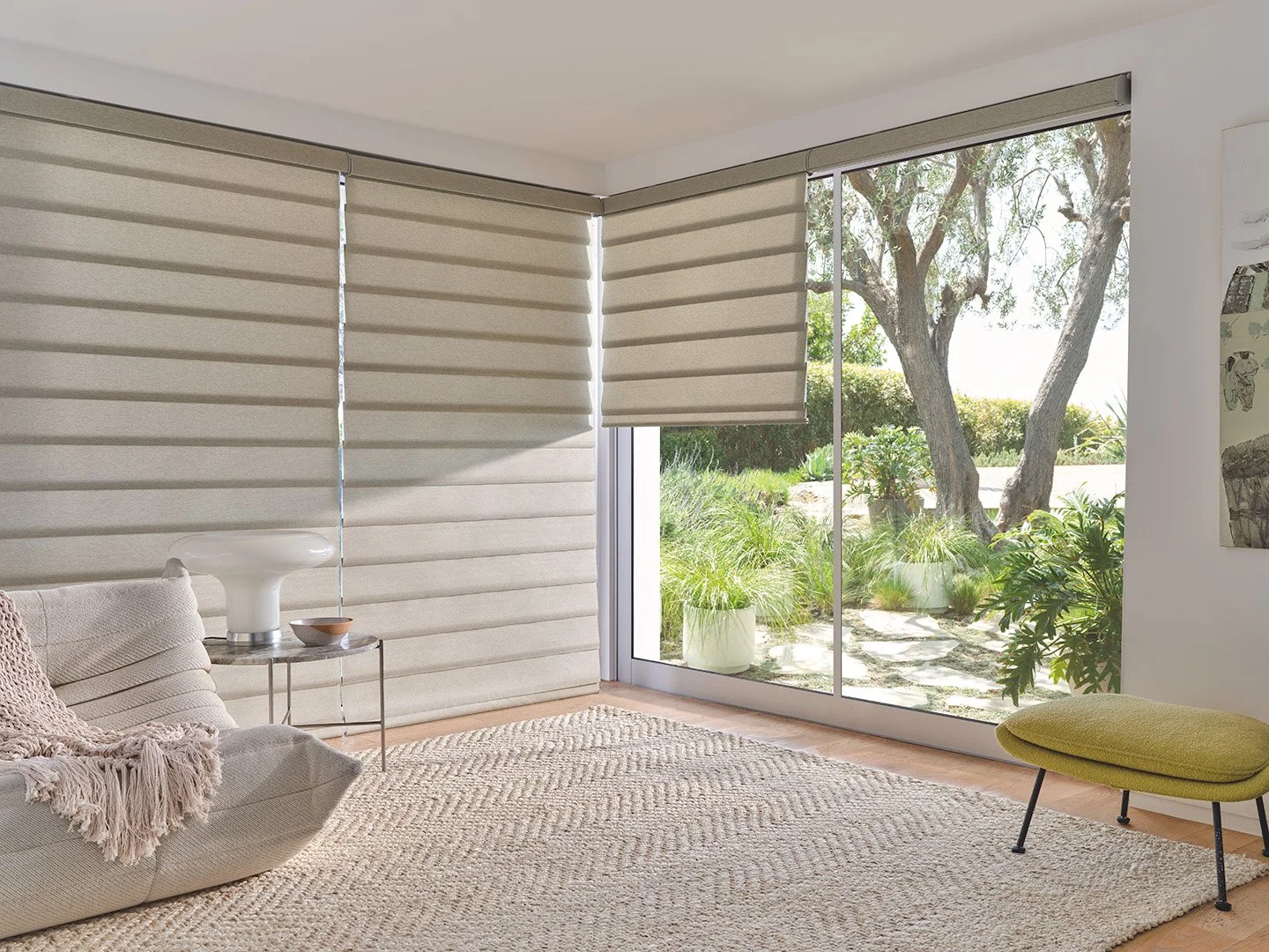 Living room with beige blinds on large sliding glass door, potted plants outside, beige sectional sofa with a blanket, small side table with a white decorative vase and bowl, woven rug, and a green ottoman.