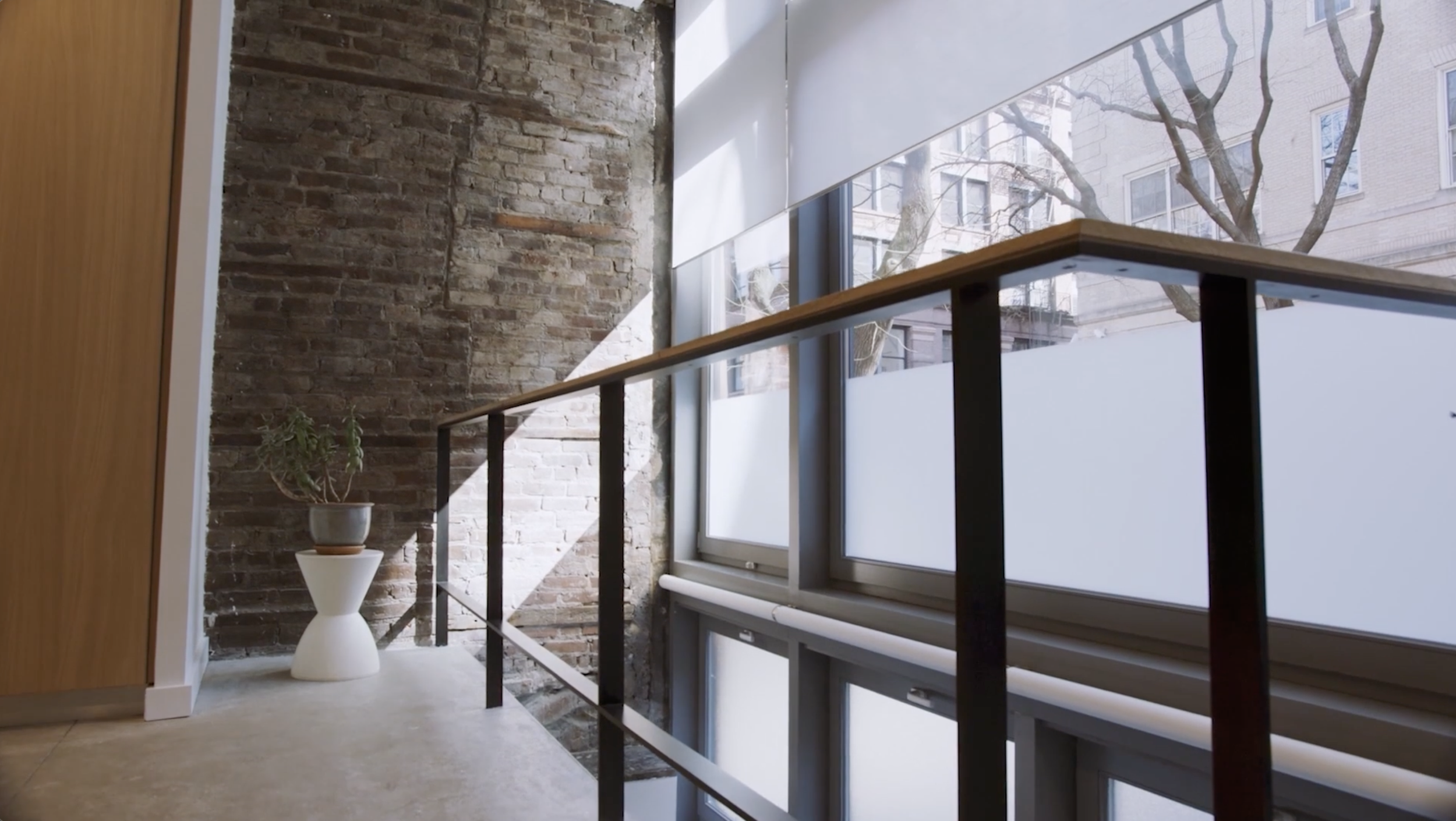 Interior space with large windows, a brick wall, a wooden panel, a white table with a potted plant, and a black metal railing.