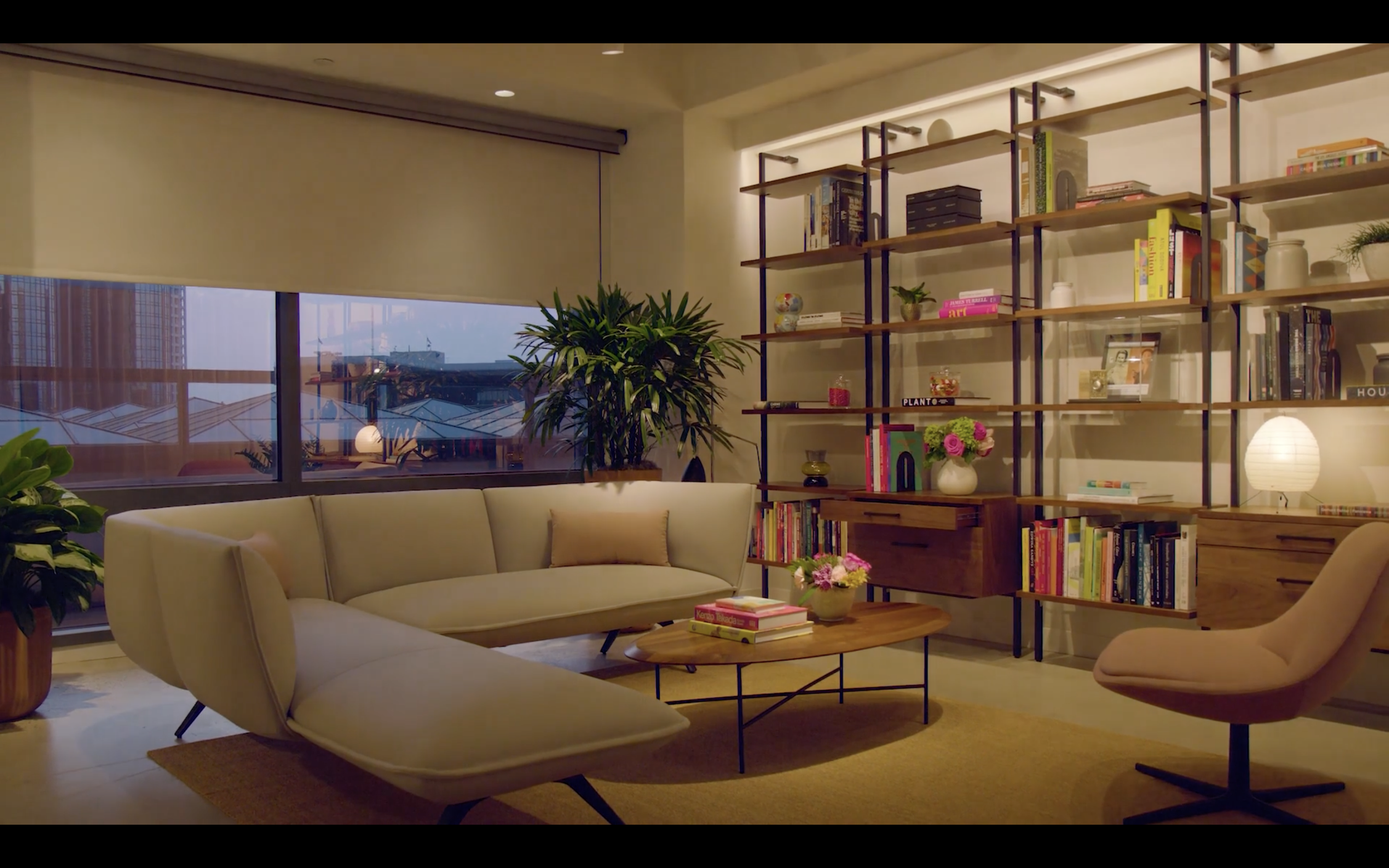 Living room with a modern beige sofa, a wooden coffee table with pink flowers and books, a tall bookshelf with books, plants, and decorative items, large windows with blinds showing buildings outside, and warm ambient lighting.