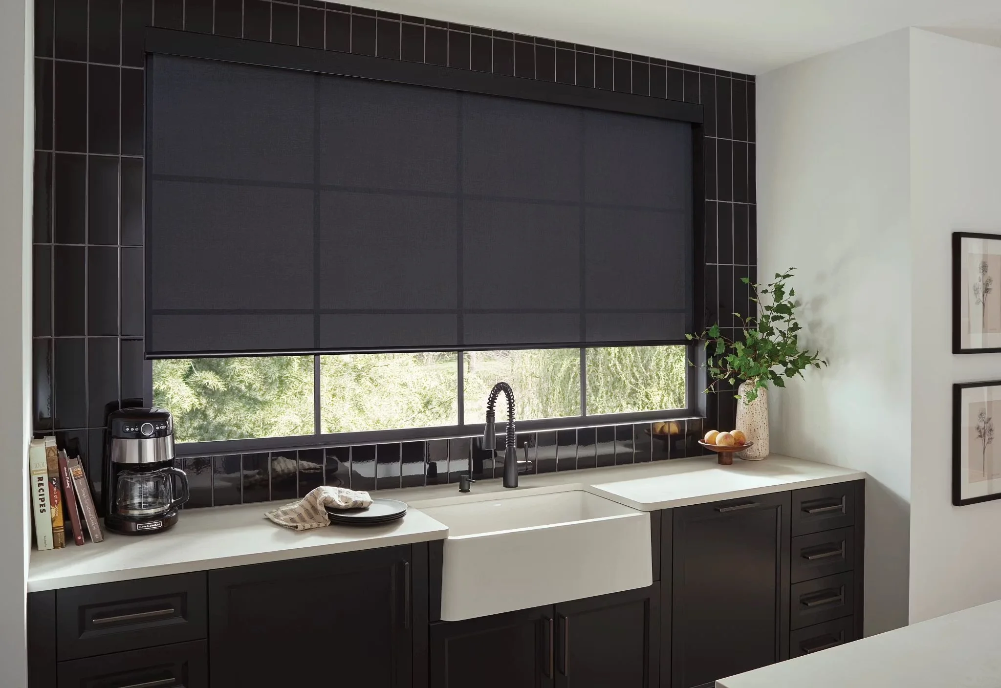 Kitchen window with black roller shade, black tiled wall, white countertop, black cabinetry, potted plant, framed artwork, coffee maker, books, plates, and a drying towel.