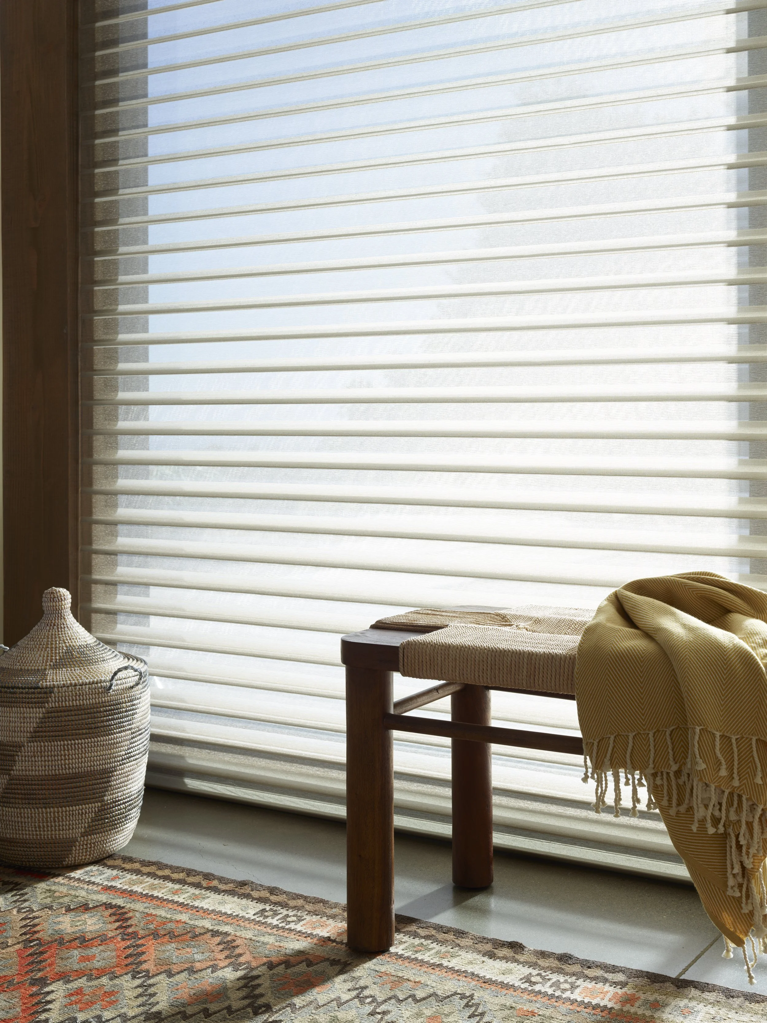 Sunlight filters through beige horizontal blinds covering a window, illuminating a corner of a room with a wooden chair with beige cushions, a round woven basket, and a patterned rug.