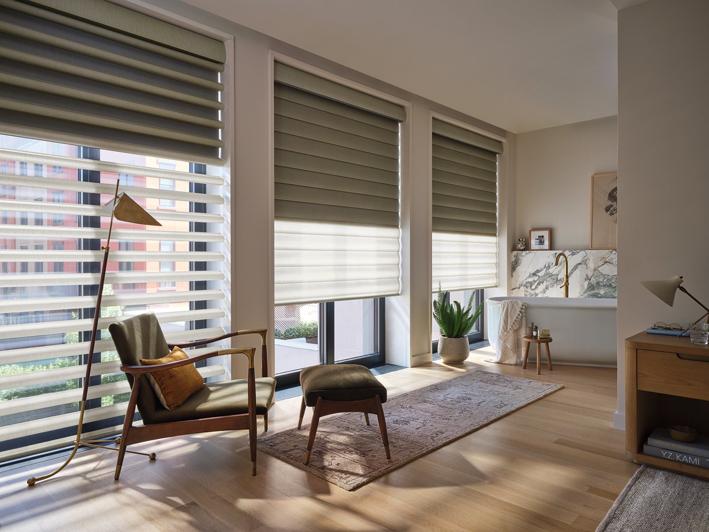 Sunlit modern living room with large windows, beige blinds, a green armchair with a brown pillow, a matching ottoman, a floor lamp, a potted plant, and a bathtub with a marble wall and a gold faucet.