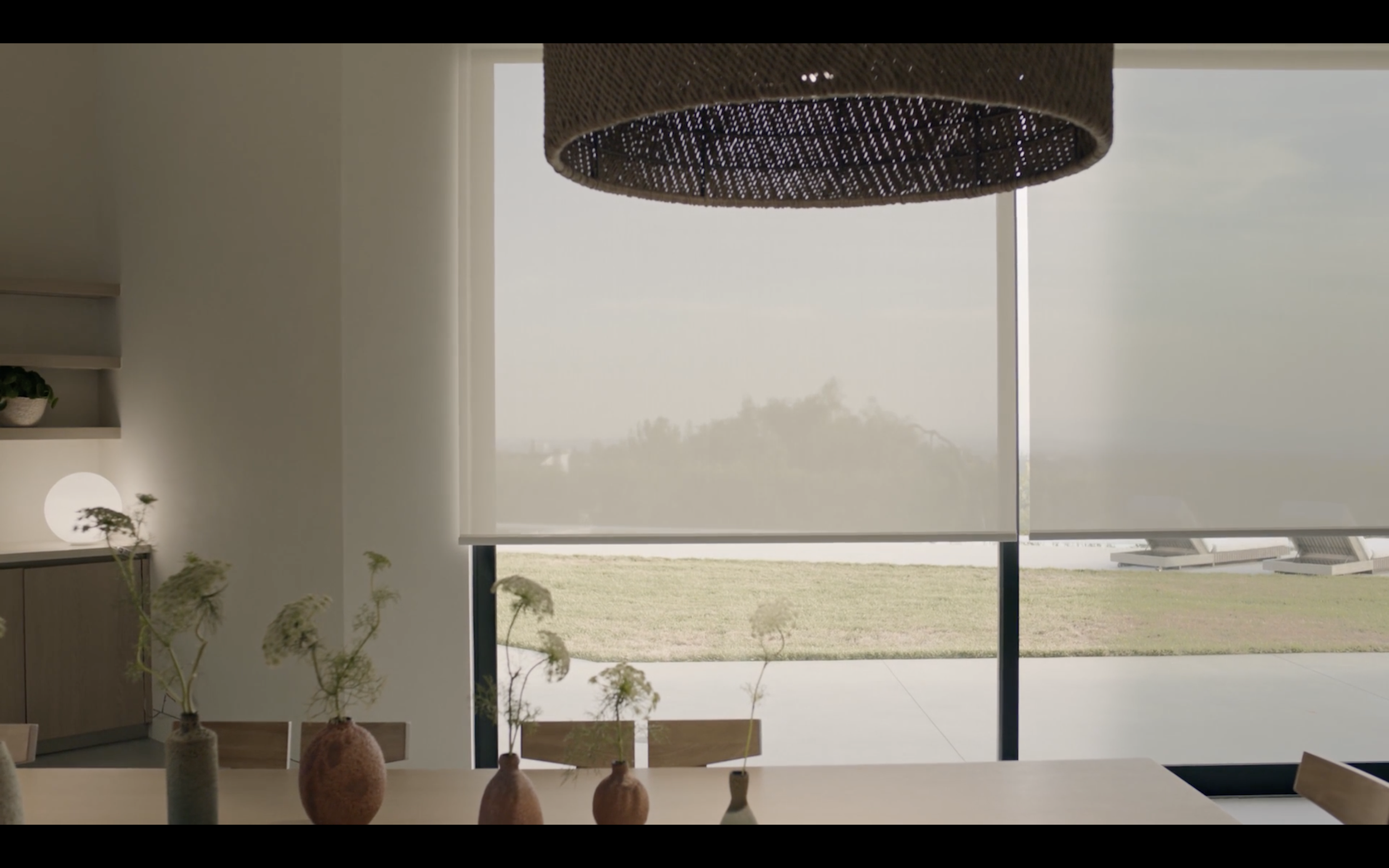 Interior view of a room with a table and decorative vases in the foreground, large window with a roller shade over a grassy outdoor area in the background.