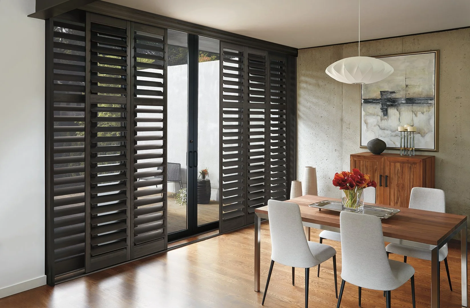Living room with a wooden dining table, four light gray chairs, a glass vase of red flowers, a wooden sideboard, abstract wall art, a pendant light, and sliding glass doors with black shutters leading to a balcony.