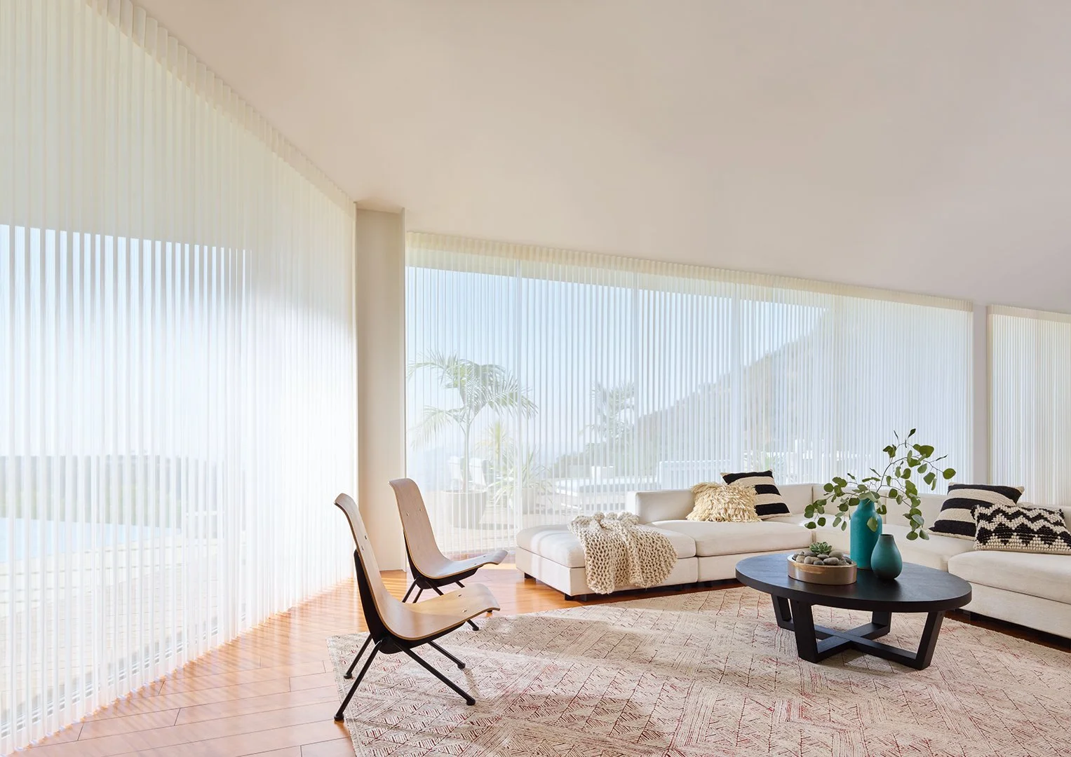 Bright, modern living room with white sectional sofa, black and white pillows, a coffee table with vases and greenery, and two wooden chairs, all in front of large windows with sheer vertical blinds and a view of outdoor palm trees.