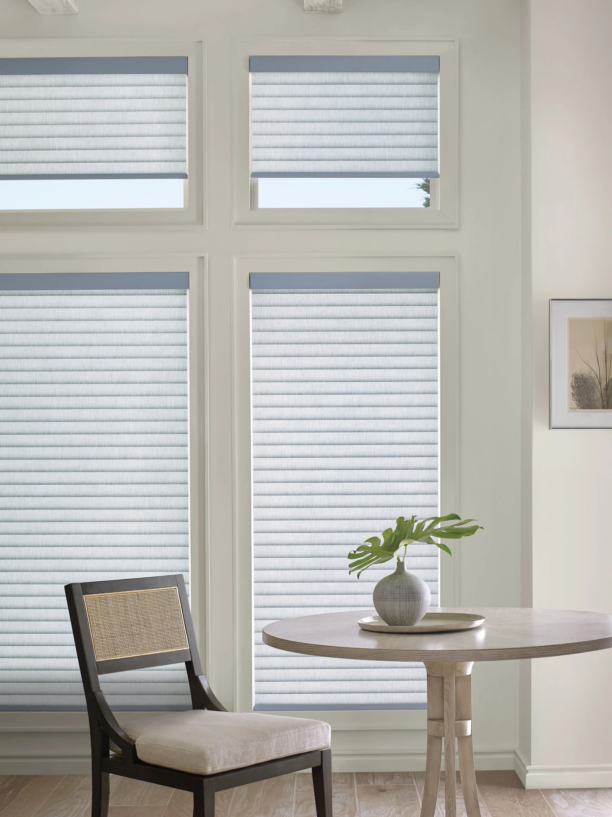 Interior view of a dining area with a round wooden table, a white vase with green monstera leaves, a black chair with a beige cushion, and four large windows with ribbed roller shades, white walls, and a framed black and white landscape photograph.