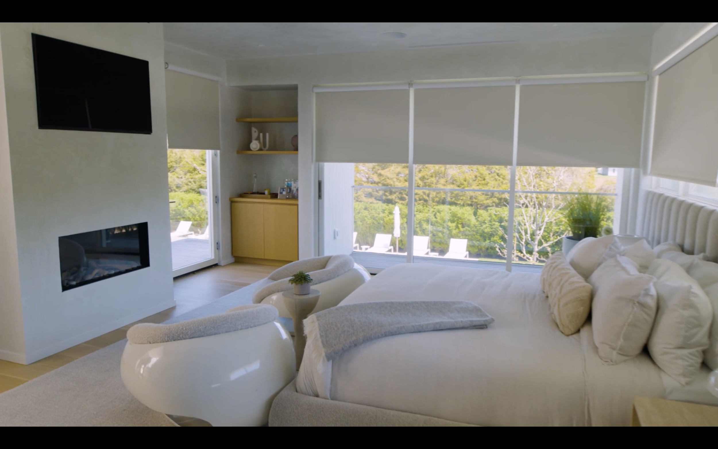 A bright living room with large windows, a white couch with beige pillows, a small plant on a side table, a white modern chair, a built-in fireplace, and a yellow cabinet against the wall, with a view of outdoor furniture and greenery outside.