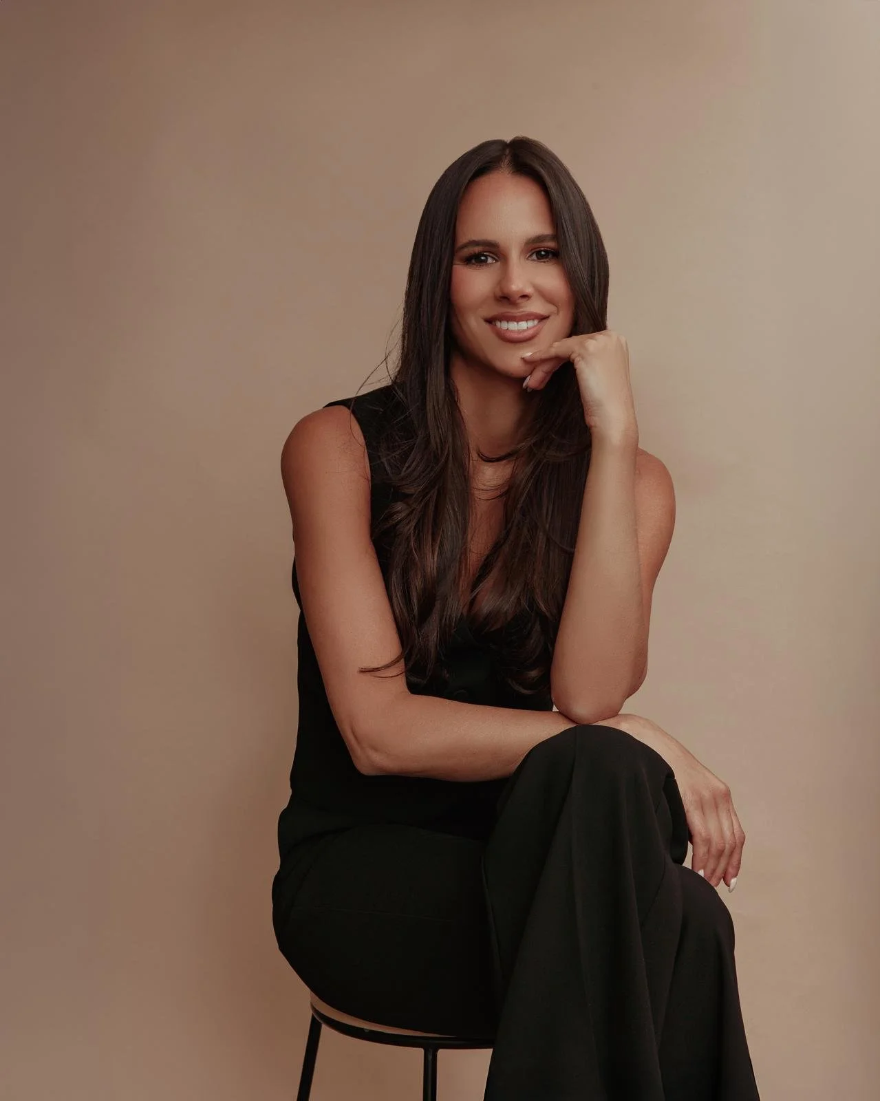 Monica Gotay dressed in black, sitting on a stool against a neutral background, smiling with her chin resting on her hand.