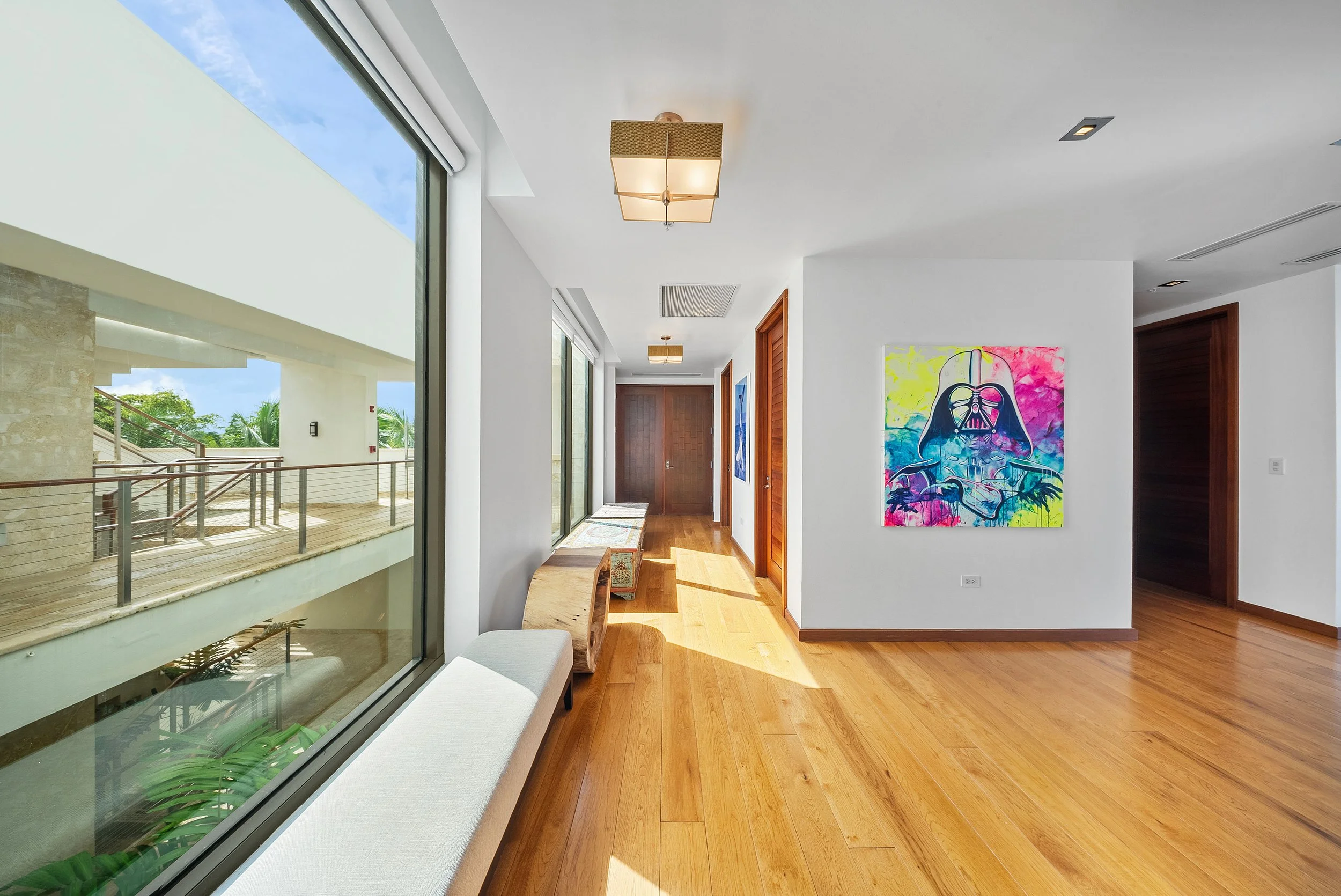 Bright corridor with wooden floors and white walls, featuring colorful art paintings and large windows allowing natural light to enter.