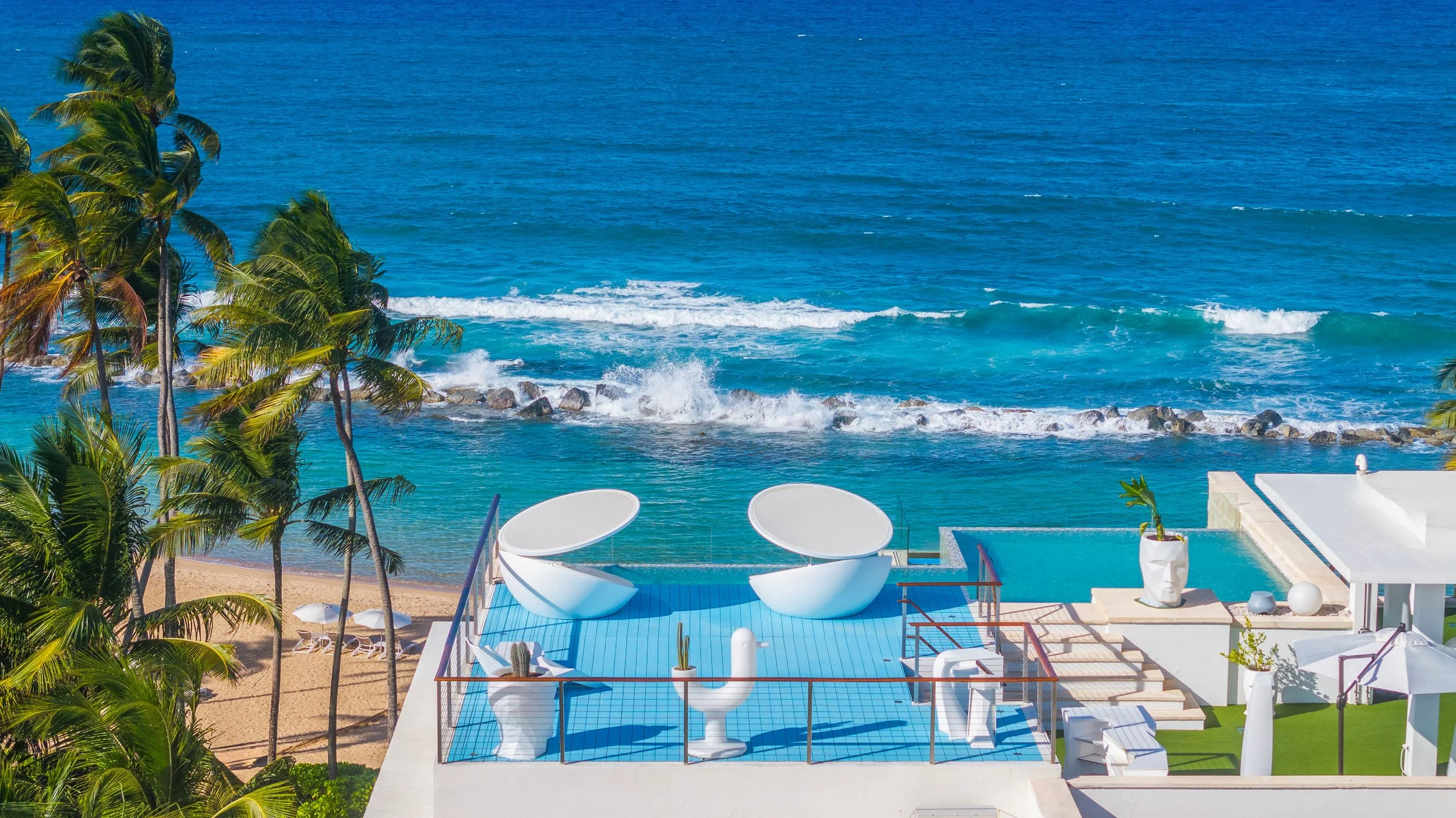 A luxurious beachside patio with white modern furniture, including two circular lounge chairs, a white sculpture, potted plants, and a pool, overlooking an ocean with palm trees, a sandy beach, and waves.