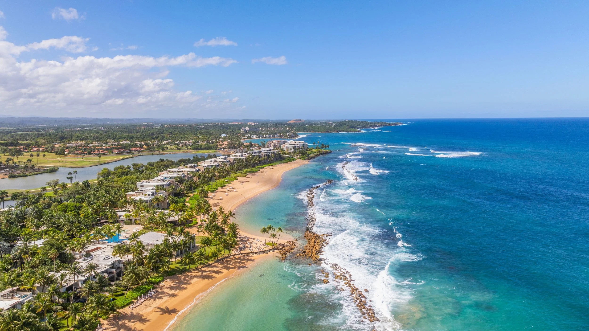 A scenic aerial view of a tropical beach with clear turquoise water, sandy shoreline, palm trees, and residential buildings along the coast under a bright blue sky with scattered clouds.