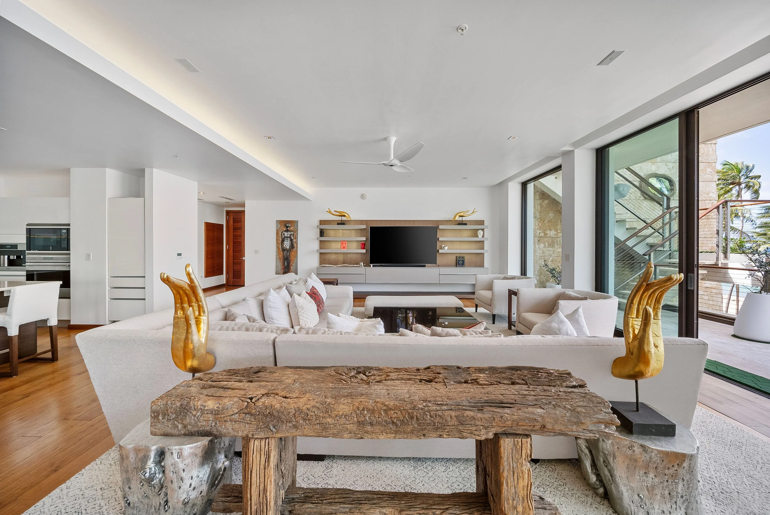 Modern living room with white sofa, armchairs, wooden coffee table, built-in shelving with television, large glass sliding doors, and decorative gold hand sculptures.