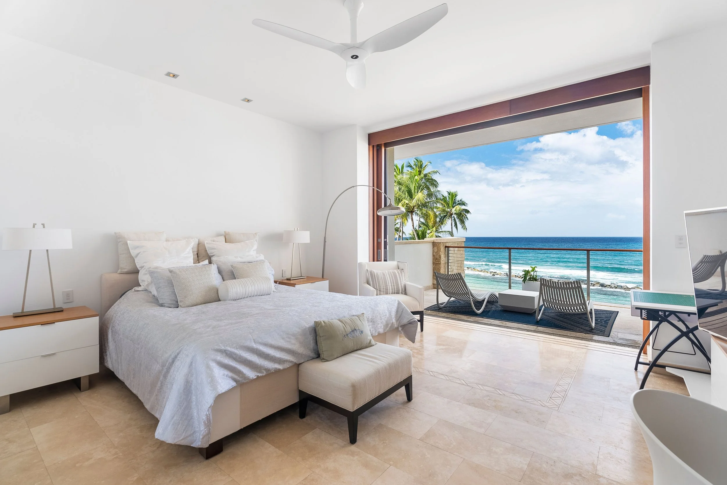A modern bedroom with a large sliding door opening to a balcony with a view of the ocean, palm trees, and blue sky.