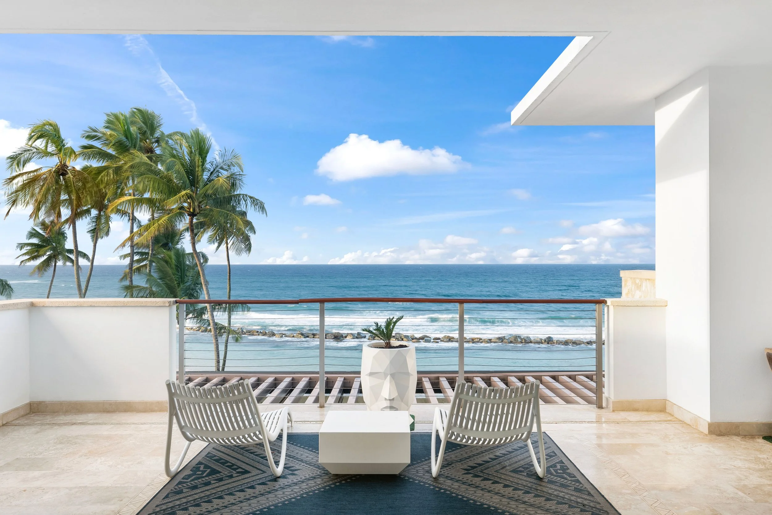 Balcony overlooking the ocean with two white chairs, a white table with a potted plant, palm trees, and a cloudy blue sky.