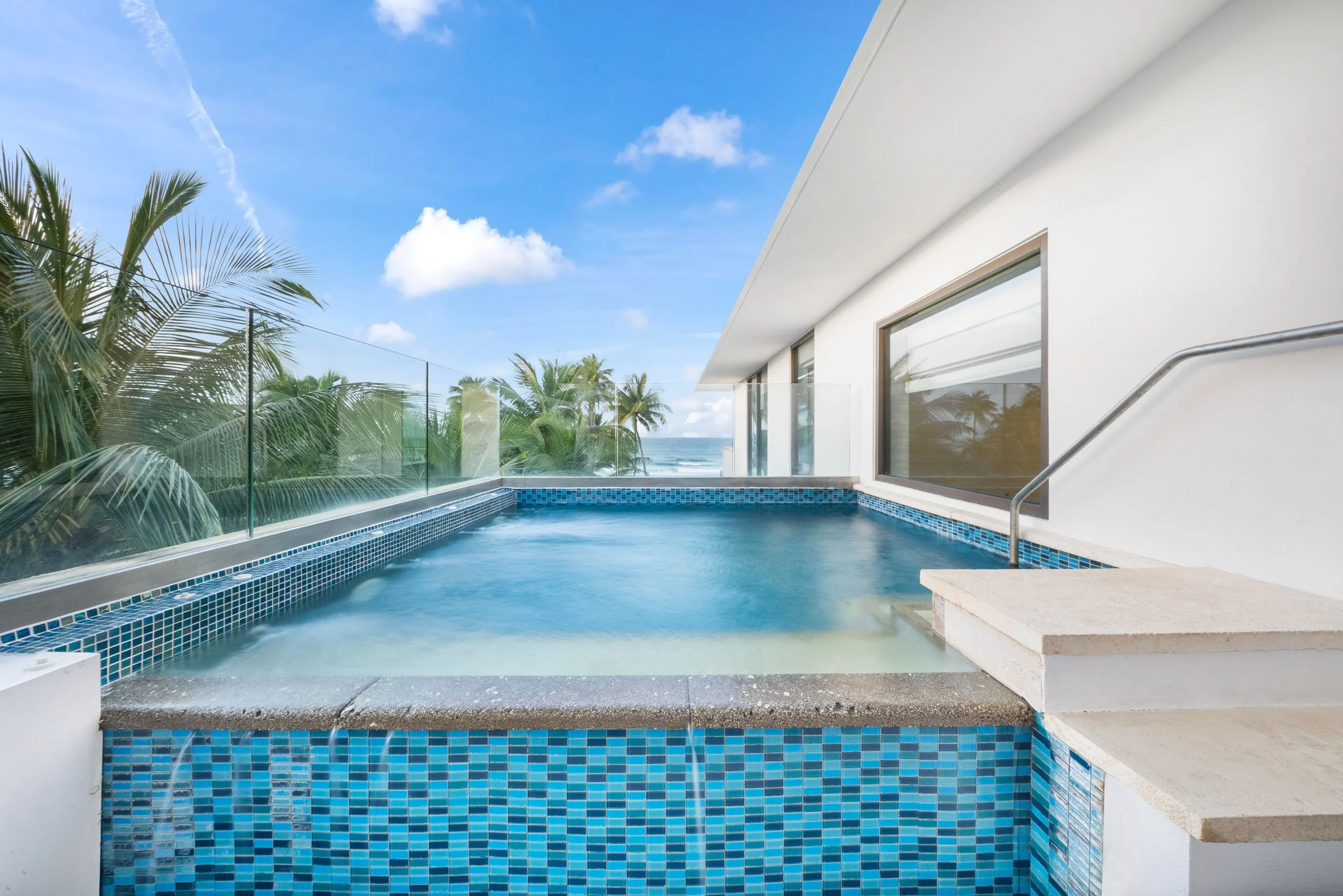 Luxury rooftop jacuzzi with view of palm trees and ocean in the background, glass railing, white building, blue sky with clouds.
