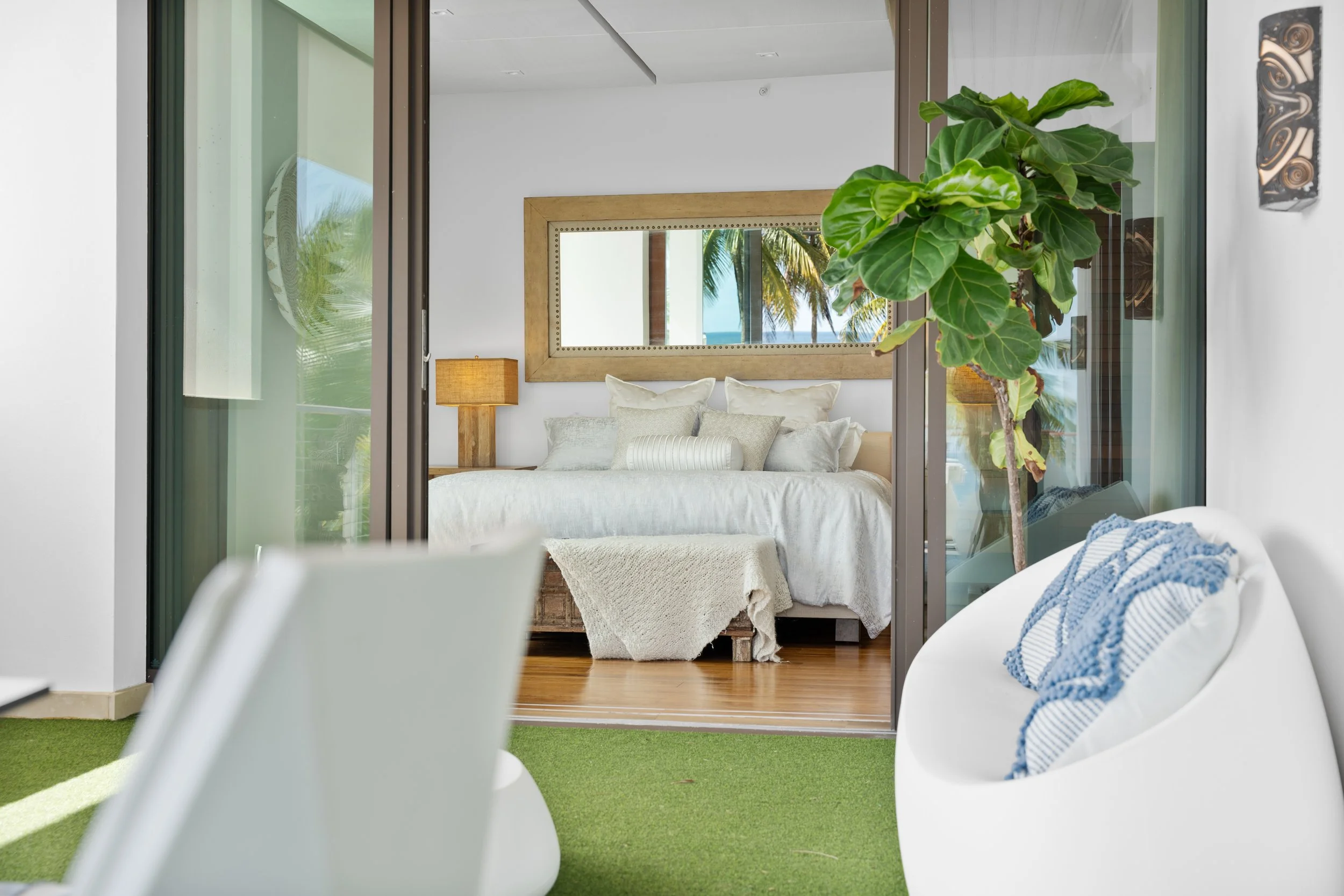 View into a bedroom through a glass sliding door, with a large bed, white linens, a mirror on the wall, a lamp, and tropical outdoor scenery visible outside.