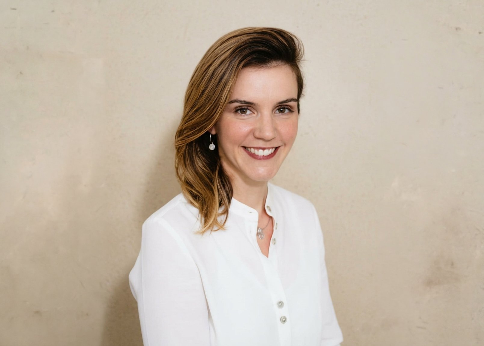 A woman with shoulder-length light brown hair, wearing a white button-up shirt and pearl earrings, smiling at the camera against a plain beige wall.