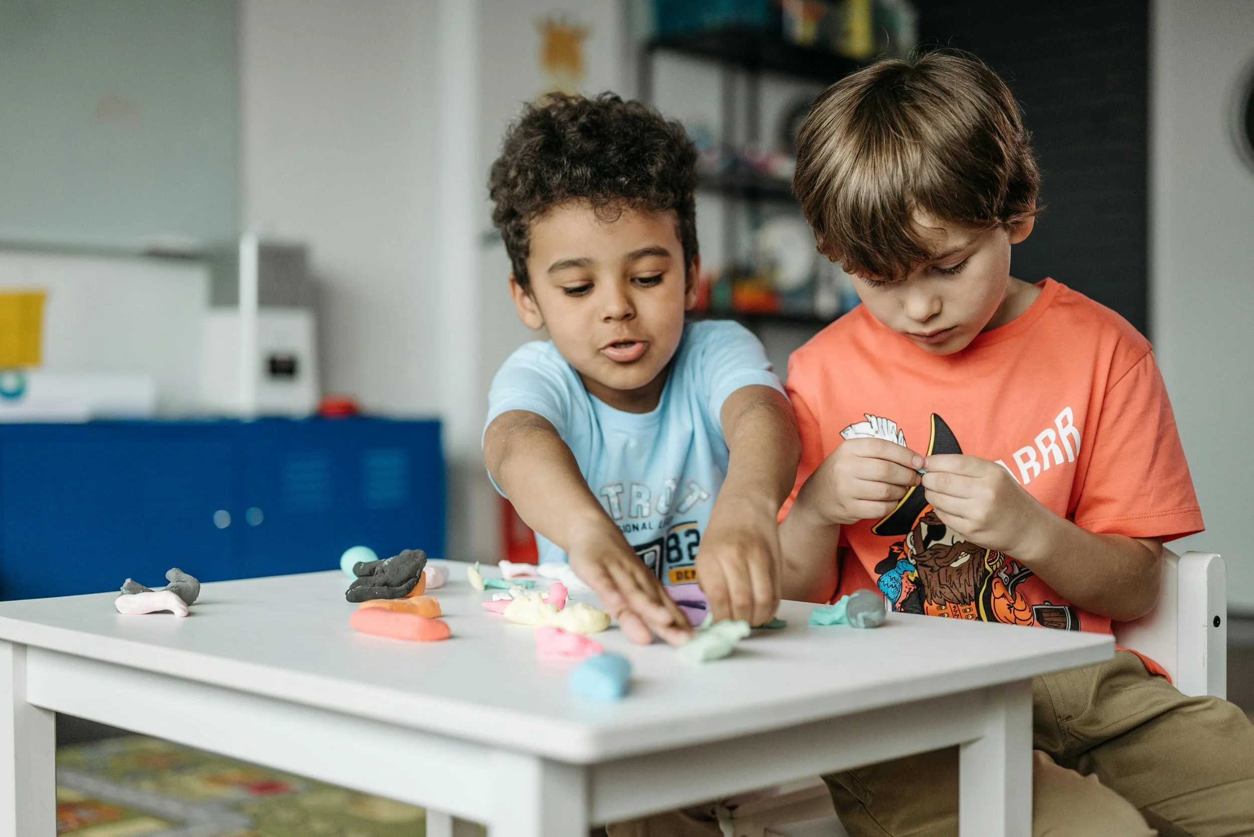 Two young boys playing with colorful clay on a white table in a playroom.